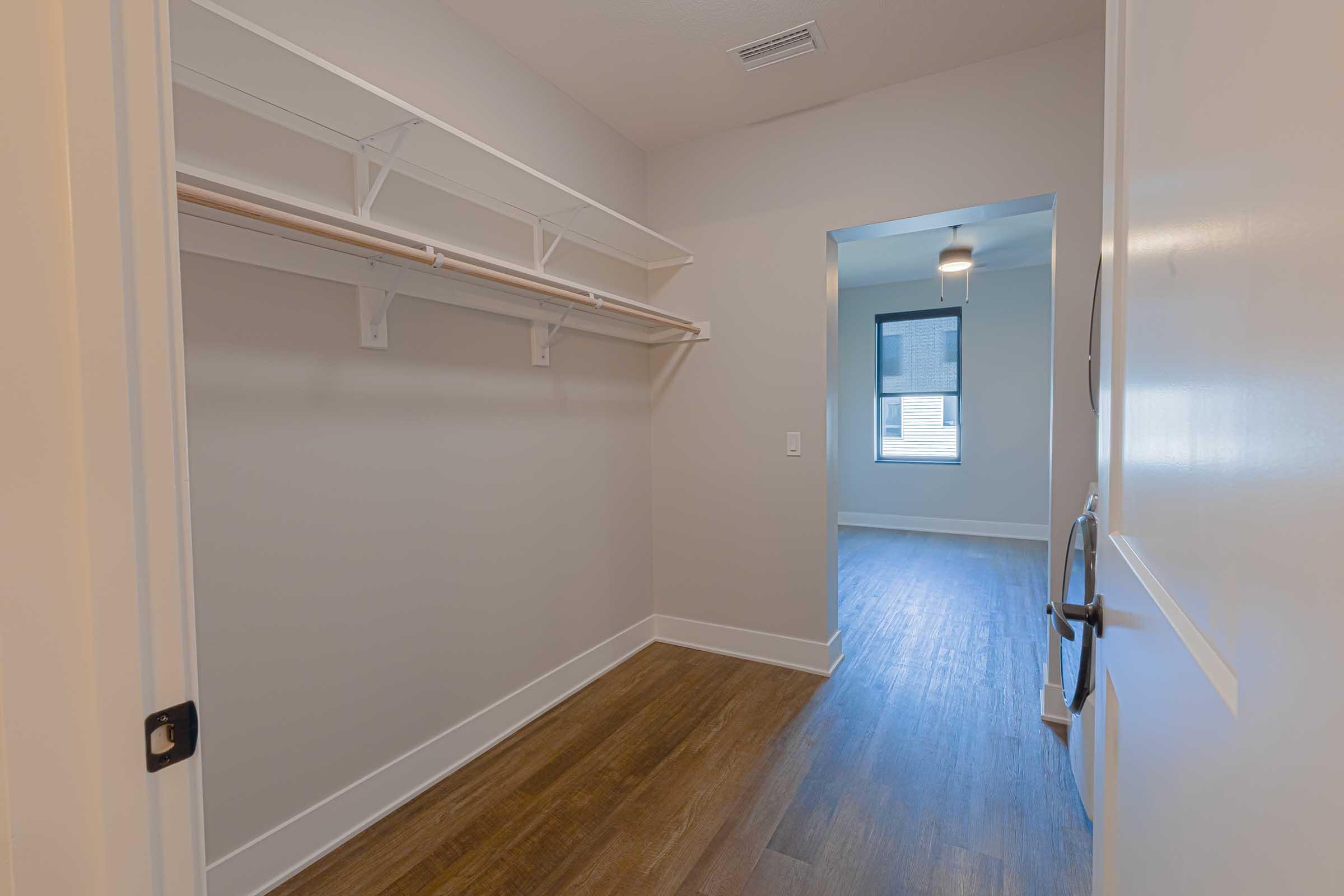 A spacious walk-in closet with white shelving and a wooden floor, leading to a well-lit room with a ceiling fan. The walls are painted in a light color, and a window with blinds is visible. The overall space appears modern and well-organized.