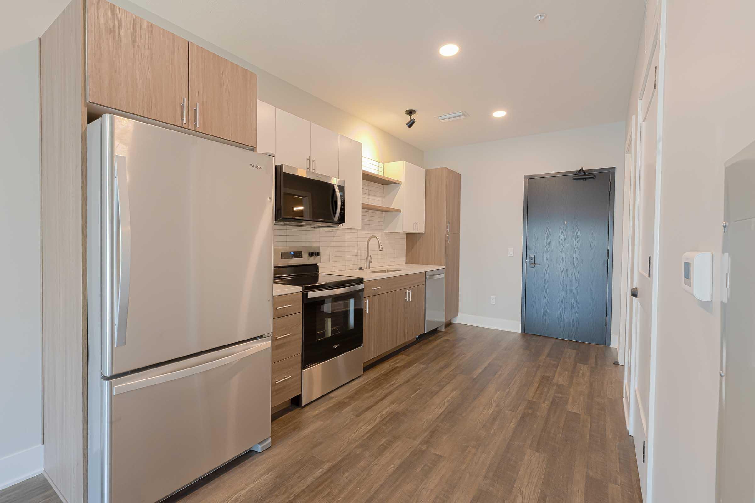 Modern kitchen featuring stainless steel appliances, including a refrigerator, oven, and microwave. The cabinets have a combination of light and dark wood finishes. The countertop is sleek and white, with a backsplash of small tiles. The floor has a warm wood-like finish, and there is a black front door visible in the background.