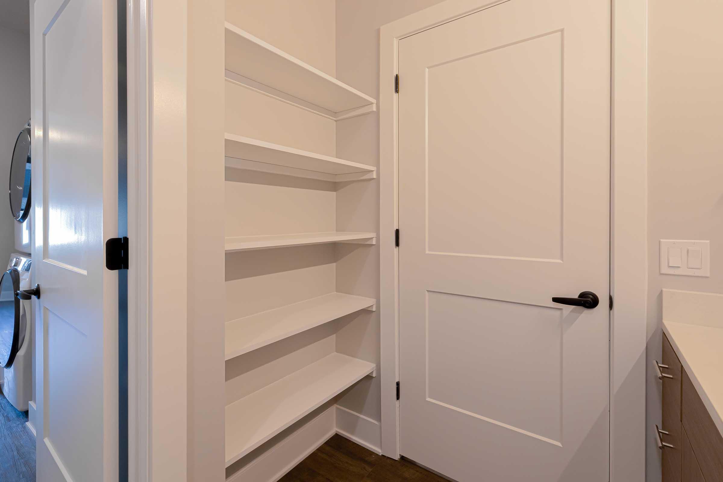 A clean, modern room featuring a white door and a wall-mounted shelf with multiple open shelves. The shelves are empty, providing a minimalist look, and the walls are painted in a light color, enhancing the spacious feel of the area. The floor is hardwood, adding warmth to the design.