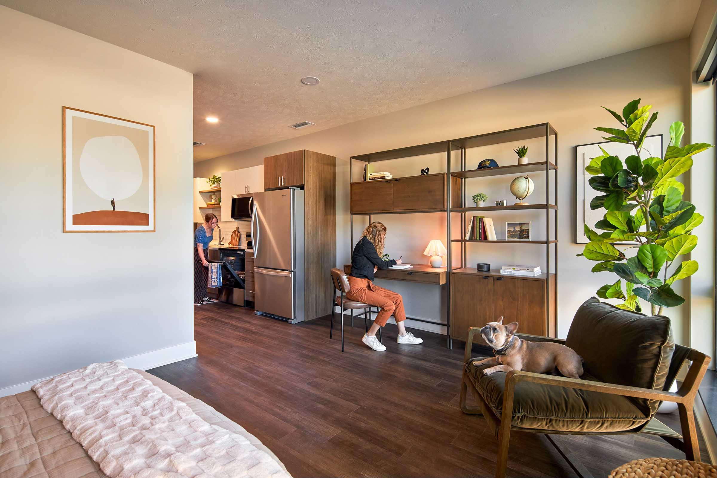 A modern, stylish apartment interior featuring a living area with a cozy chair and a dog resting, a kitchen in the background with a person cooking, and another person working at a desk. The decor includes plants and framed artwork, contributing to a warm and inviting atmosphere.