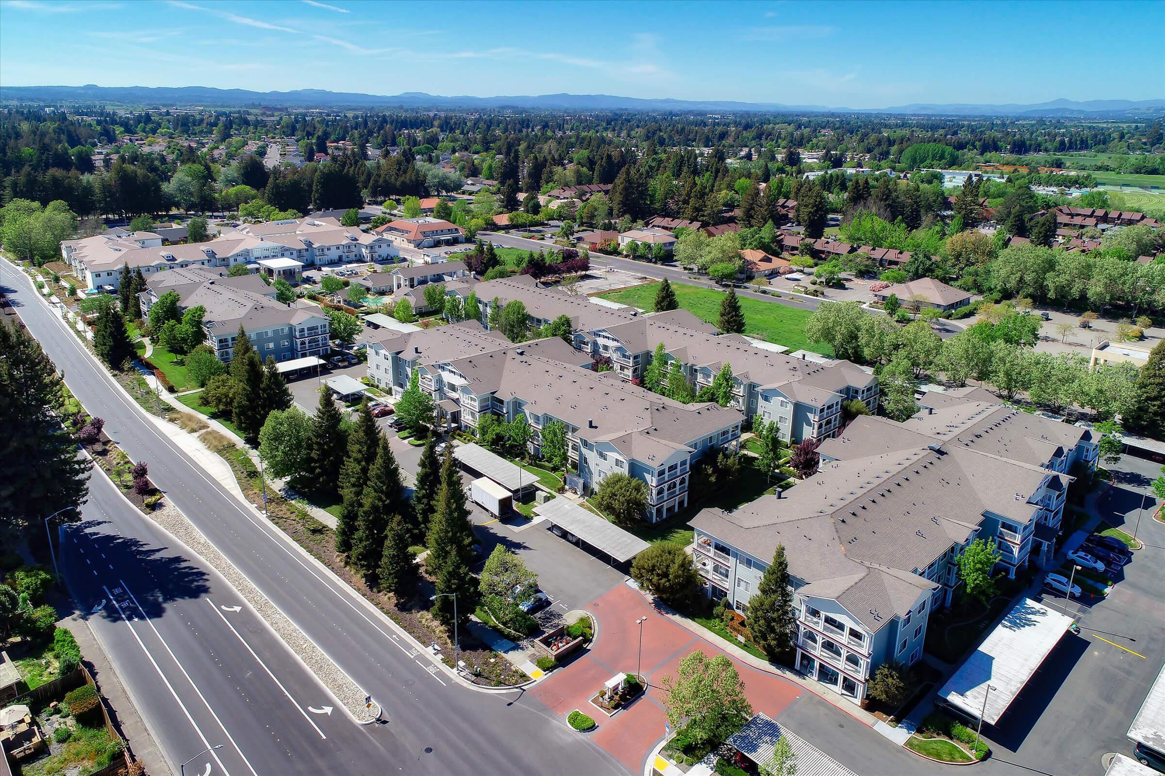 Aerial view of a suburban neighborhood featuring residential buildings, tree-lined streets, and open green spaces. The landscape includes multiple low-rise apartment complexes and well-maintained lawns, with mountains in the distance under a clear blue sky.
