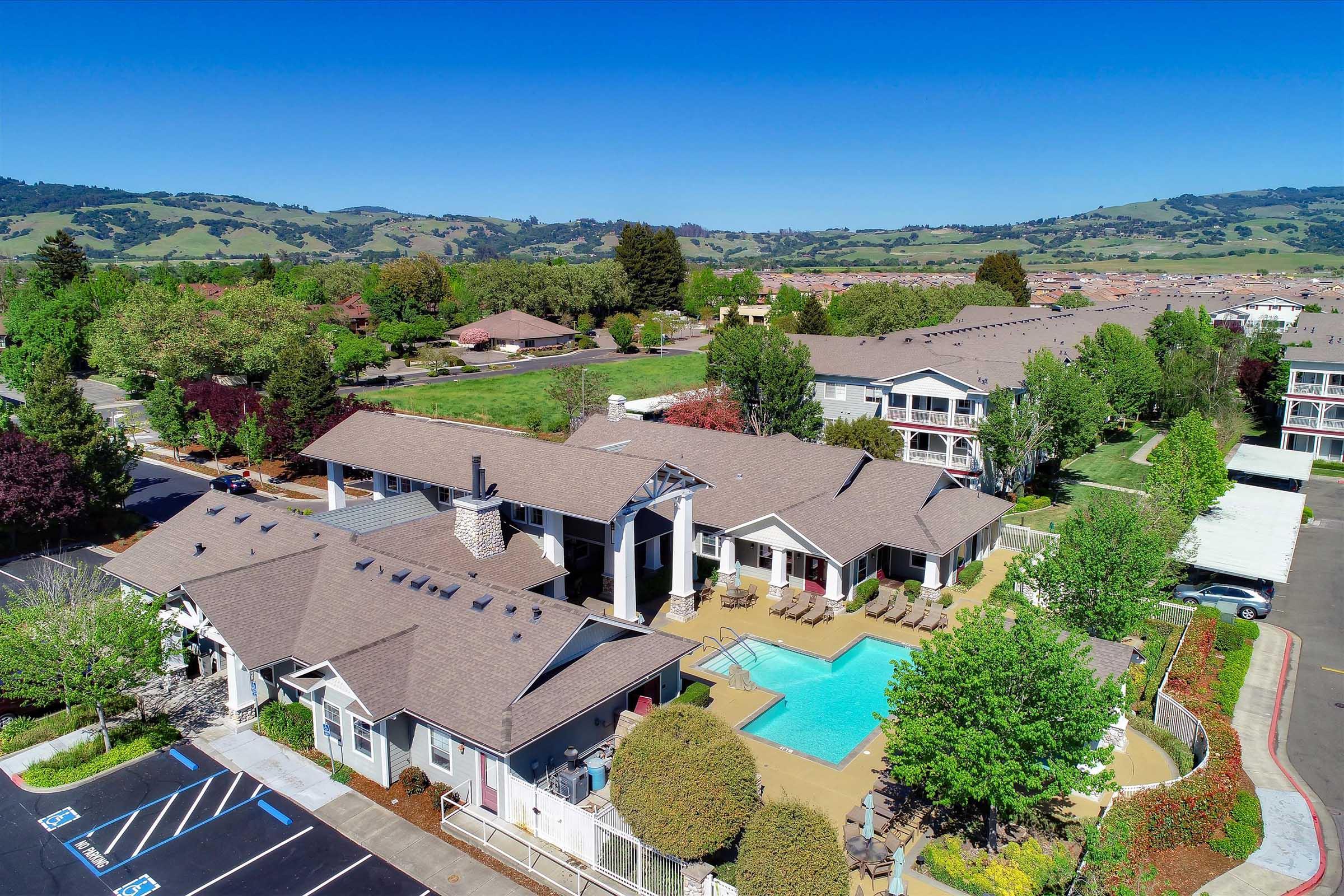 Aerial view of a residential complex featuring multiple buildings, a swimming pool surrounded by lounge chairs, and landscaped greenery. The background includes rolling hills and clear blue skies, showcasing a picturesque suburban setting. Parking spaces are visible in the foreground.