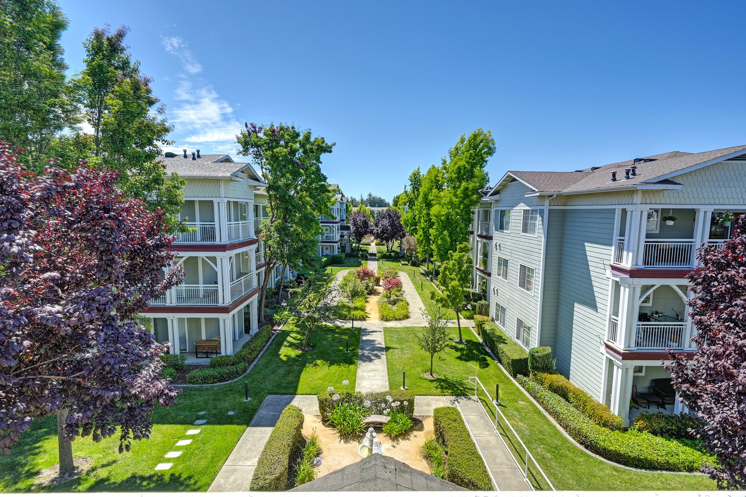 View of a residential complex featuring well-maintained landscaping with green lawns, flowering plants, and trees. The buildings are two to three stories high, surrounded by pathways and communal areas, all under a clear blue sky.