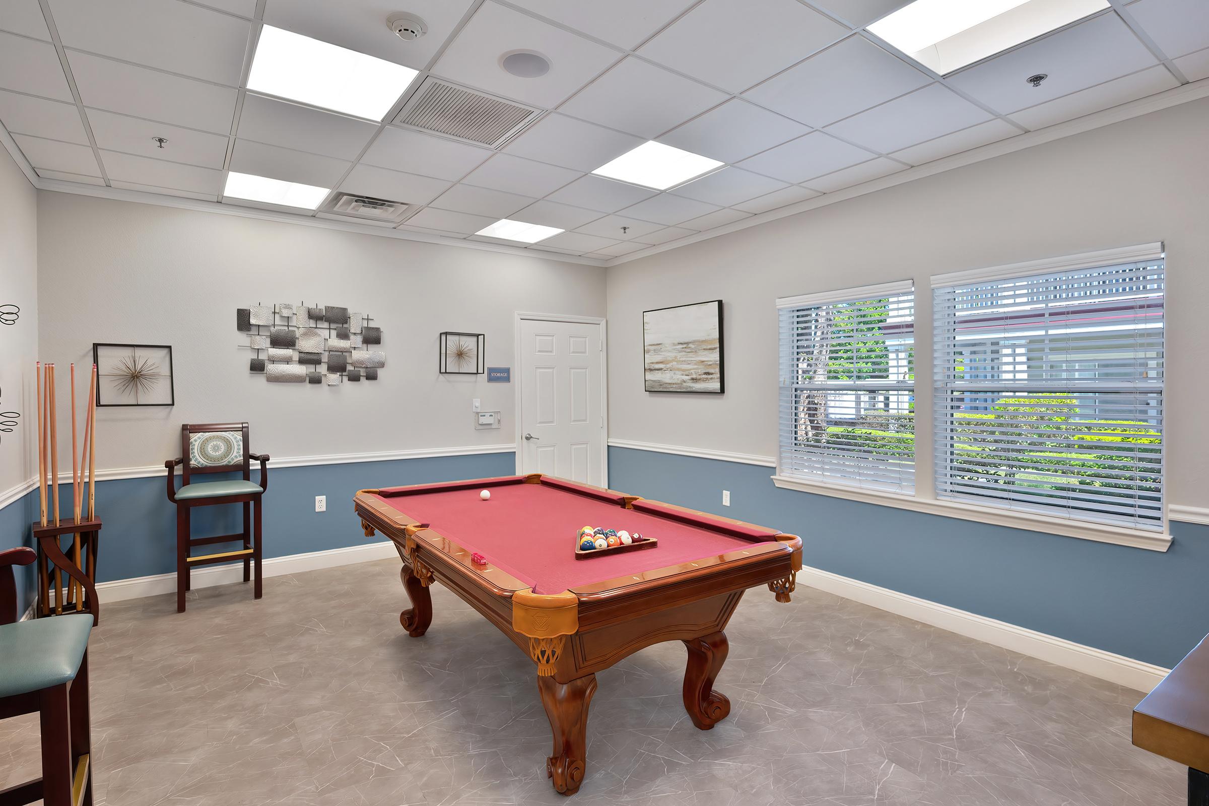 A bright room featuring a red pool table at the center. Surrounding walls are decorated with framed art, and there's a window with blinds allowing natural light. A small chair and table are positioned in one corner. The floor is a light gray tile, enhancing the spacious feel of the area.