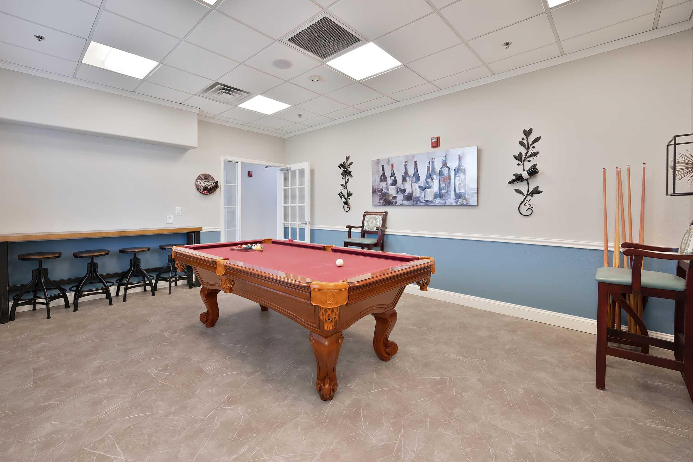 A well-lit game room featuring a polished billiards table with colored balls arranged on it. The walls are decorated with artwork, and there's a bar area with high stools. The flooring is light-colored, and there are a few chairs nearby.