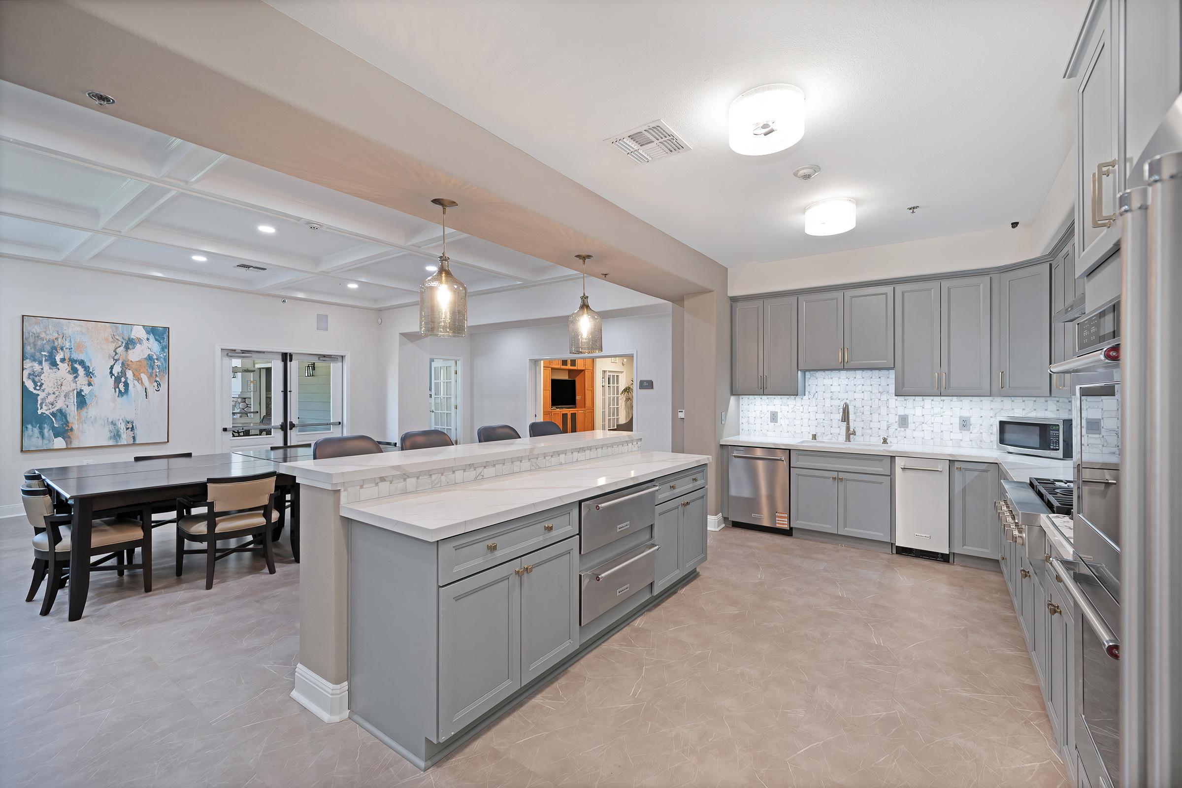 A spacious modern kitchen with gray cabinetry, a large island with a white countertop, stainless steel appliances, and pendant lighting. A dining area with a table and chairs is visible in the background, along with a large artwork on the wall, creating a bright and inviting atmosphere.