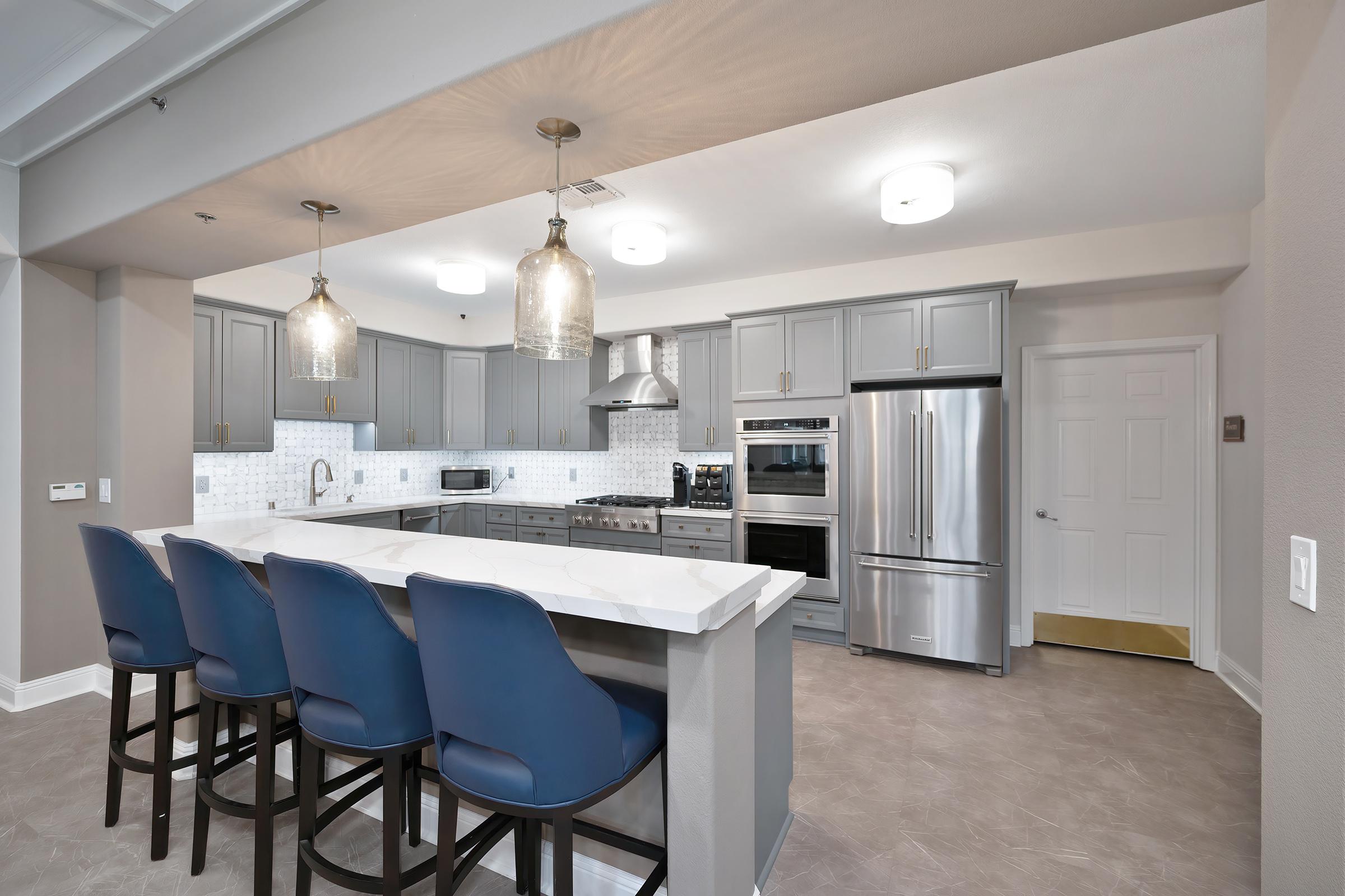 Modern kitchen featuring gray cabinets and a marble island with four blue bar stools. Equipped with stainless steel appliances, including an oven, microwave, and refrigerator. Pendant lighting hangs above the island, and a white tile backsplash complements the overall design. Walls painted in neutral tones.
