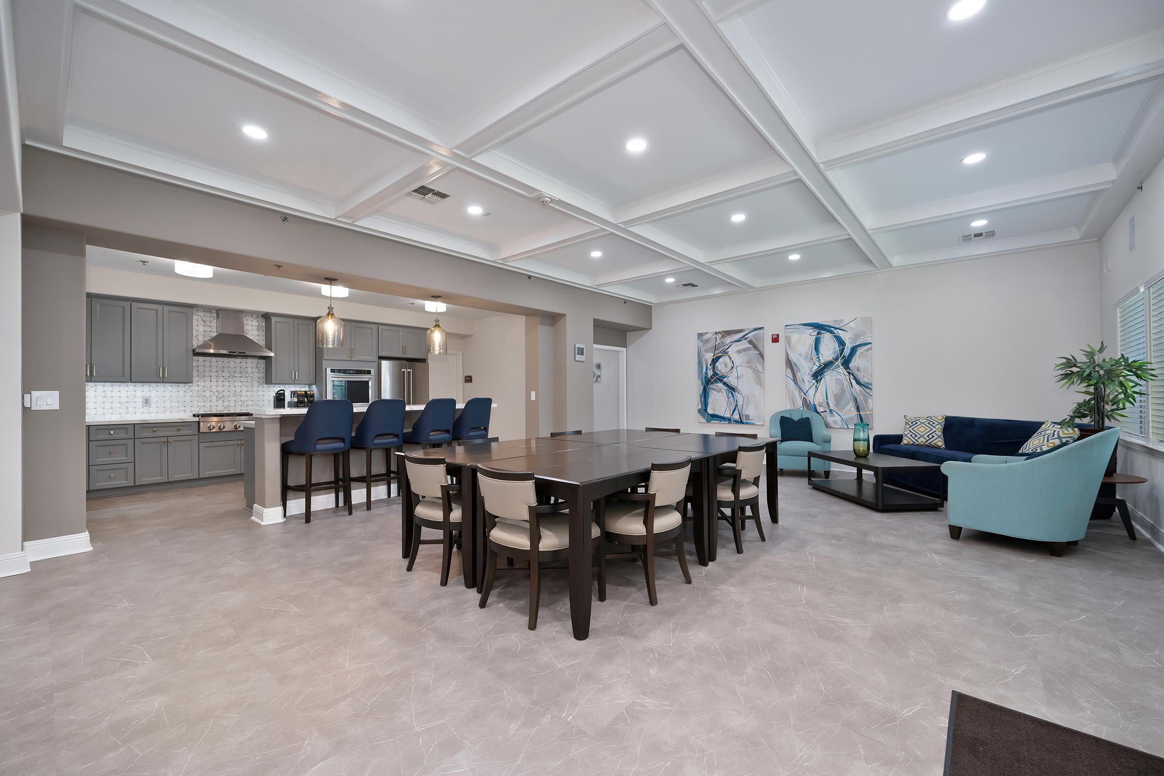 A spacious modern room featuring a dining area with a large table and chairs, a living area with a blue couch and armchair, and a kitchen in the background. The walls are painted in neutral tones, and there are abstract artworks on the wall, along with bright natural light from the windows.