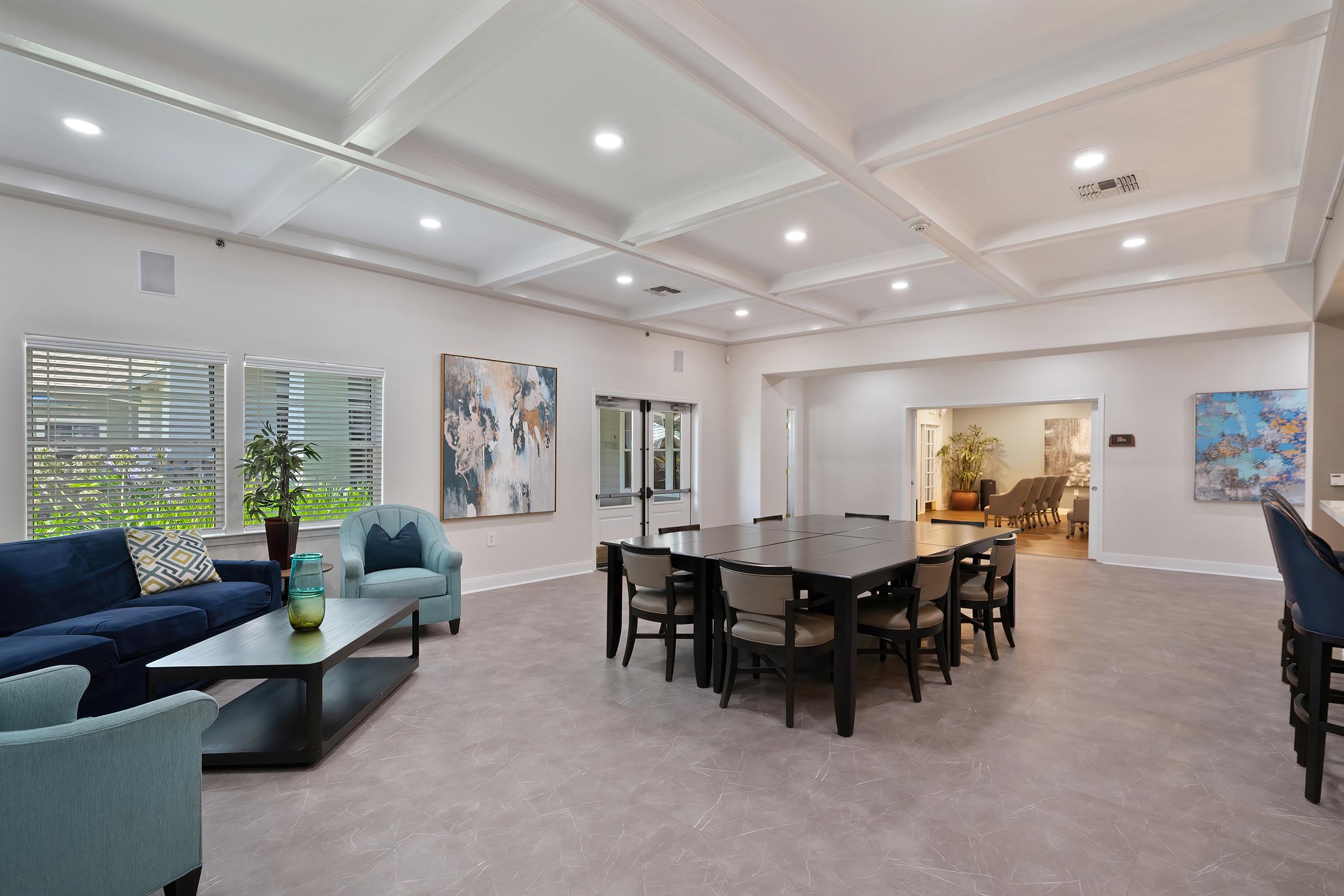 A bright, spacious interior of a modern living area featuring a blue sofa, a coffee table, and a dining table with chairs. The room has coffered ceilings, recessed lighting, and large windows, creating a welcoming atmosphere. Artwork adorns the walls, and plants add a touch of greenery.