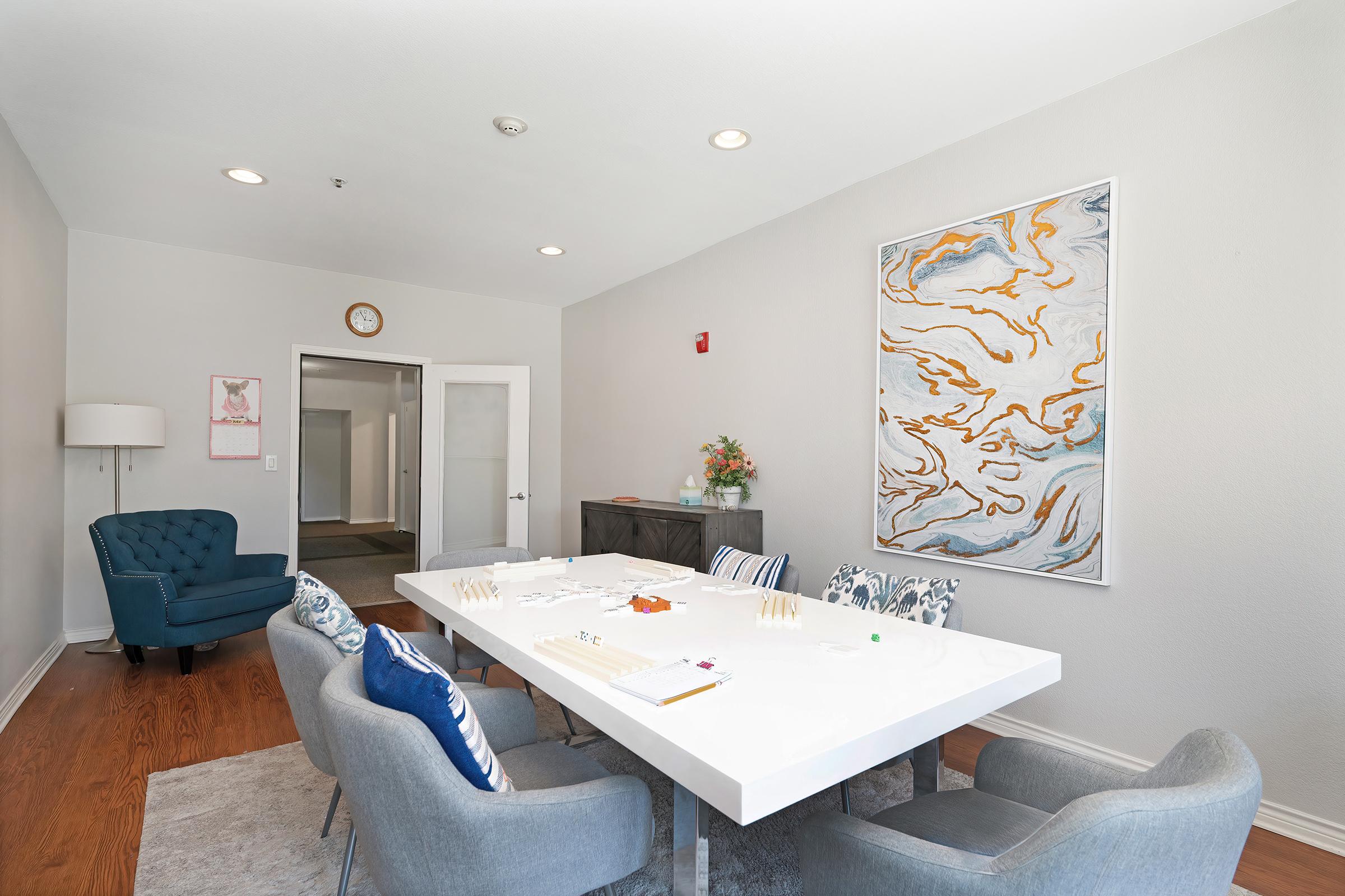 A modern meeting room featuring a large white table surrounded by blue and gray chairs. A piece of abstract art hangs on the wall, and there's a lamp in one corner. The room has natural light and a neutral color palette, creating a clean and inviting space.