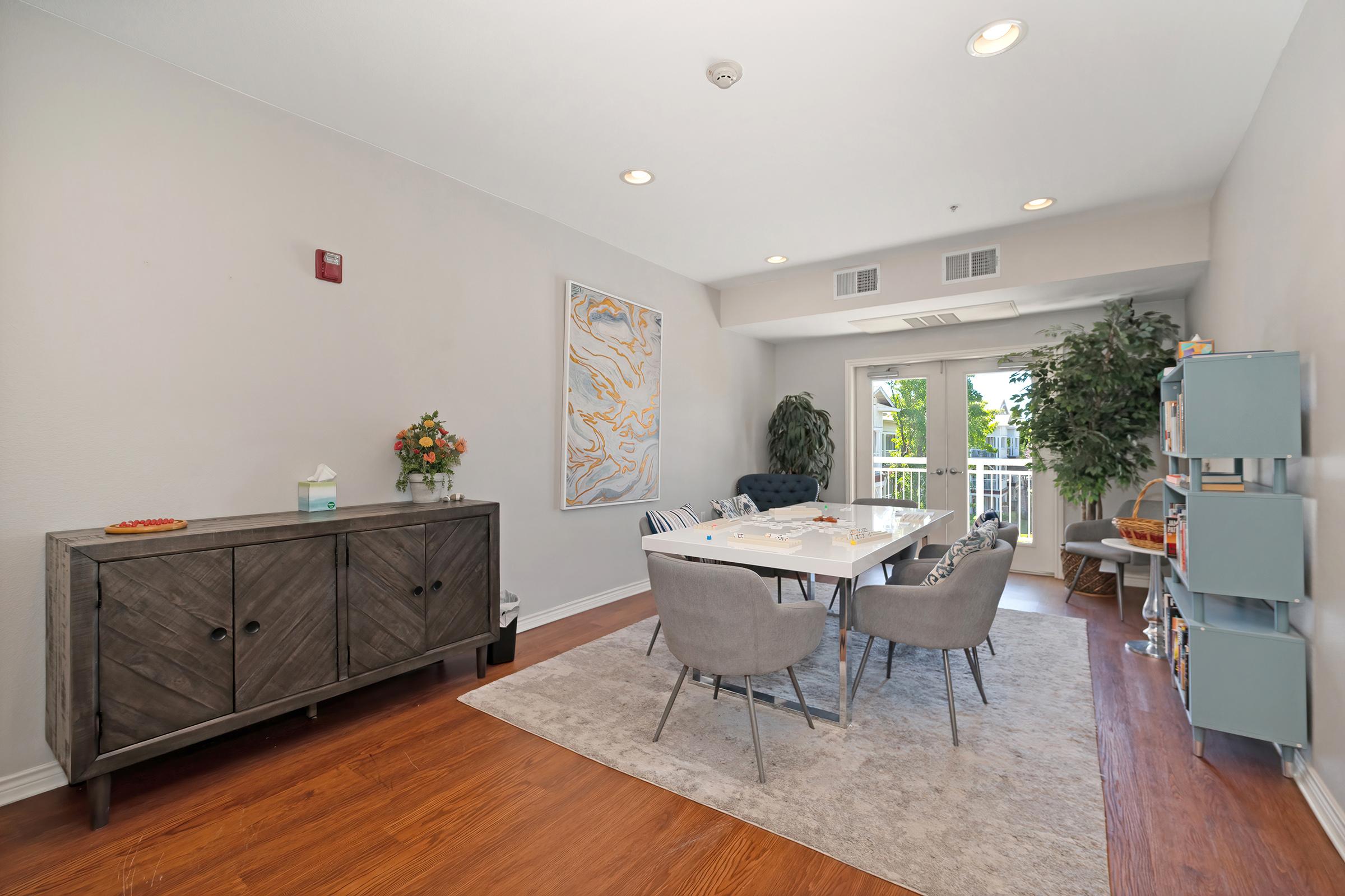 A modern dining area featuring a light wood table surrounded by gray upholstered chairs. There's a dark wooden sideboard against the wall, a decorative vase with flowers, and abstract art on the walls. Large windows let in natural light, and greenery decorates the space, creating a welcoming atmosphere.