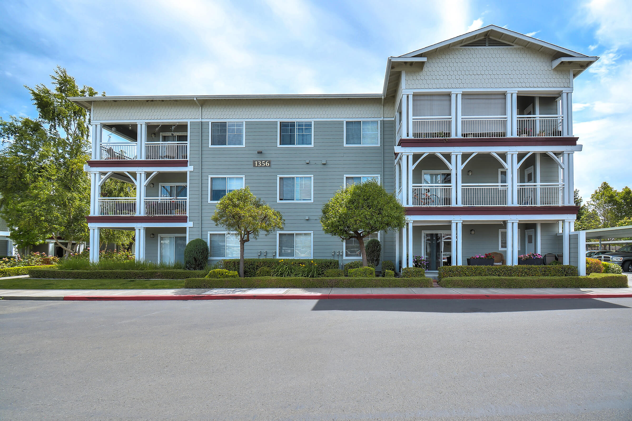 A modern multi-story apartment building featuring a light blue exterior with white trim. The building has several balconies, neatly landscaped shrubs, and trees in front. The street is visible in the foreground, with a clear blue sky above.