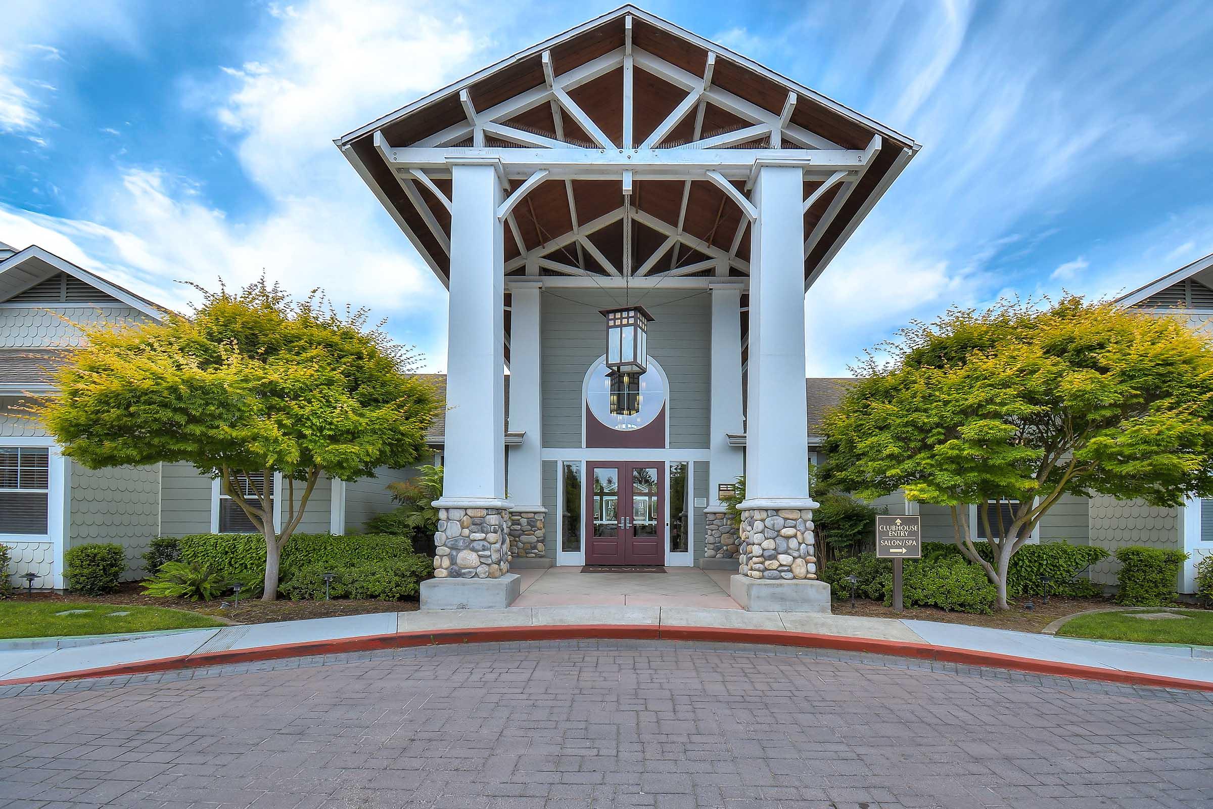A welcoming building entrance with a peaked roof and decorative pillars. Flanked by neatly trimmed trees and shrubs, the front features a large circular window and a double door entryway. The pathway is paved, and the sky above is partly cloudy, adding to the inviting atmosphere.