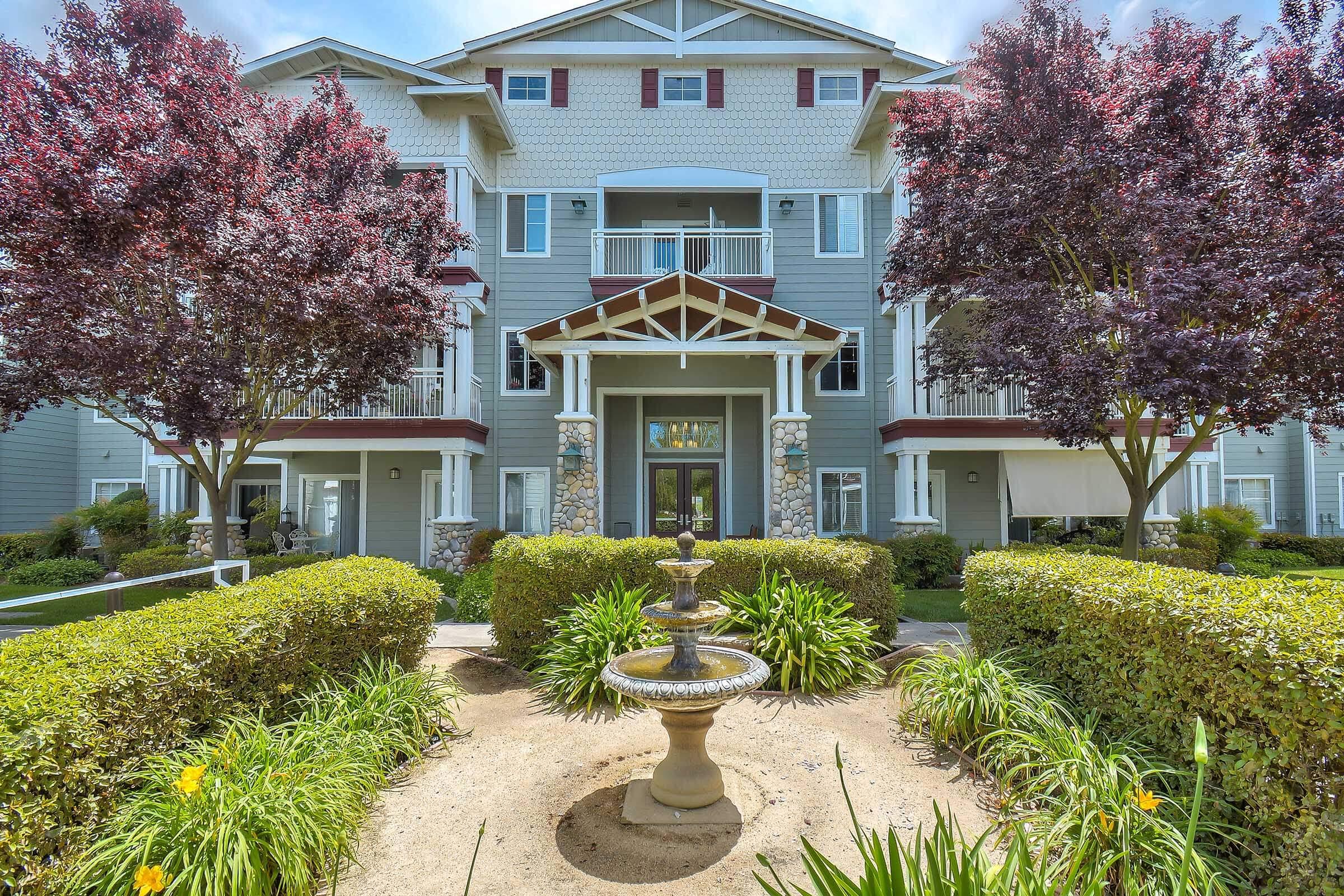 Exterior view of a multi-story apartment building featuring balconies and a central entrance. The facade is decorated with a mix of light and dark siding. A landscaped area with a fountain, vibrant green plants, and blooming flowers is in the foreground, complemented by trees with purple leaves.