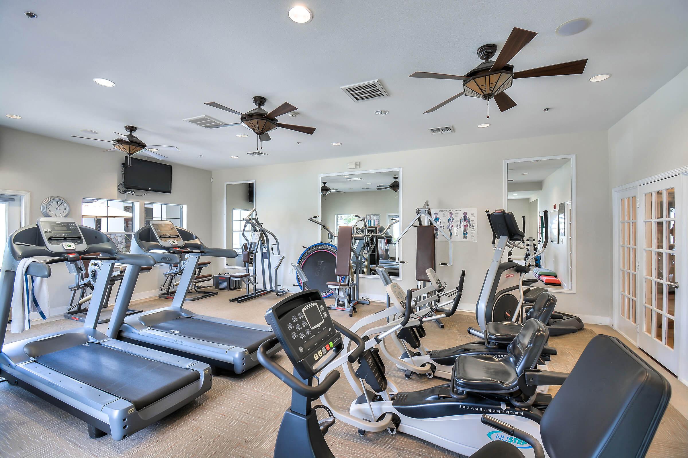 A modern gym interior featuring several exercise machines including treadmills, ellipticals, and stationary bikes. The space is well-lit with ceiling fans and has a wall-mounted TV. Large windows allow natural light, and there are fitness posters on the wall.