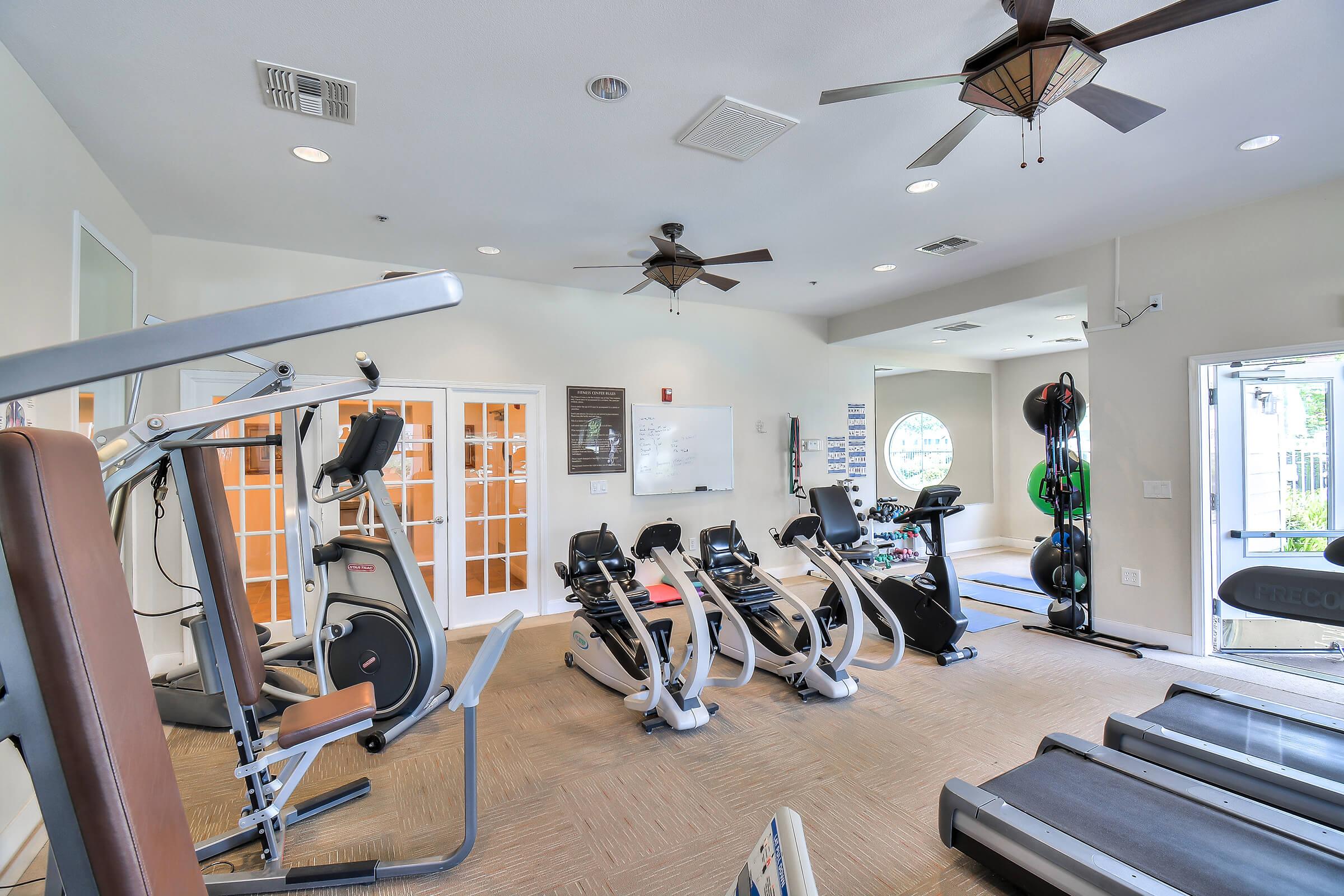 A bright and spacious gym featuring several exercise machines, including treadmills, stationary bikes, and rowing machines. The room has ceiling fans, large windows allowing natural light, and a whiteboard for workouts. The decor is modern and inviting, with a clean and organized appearance.