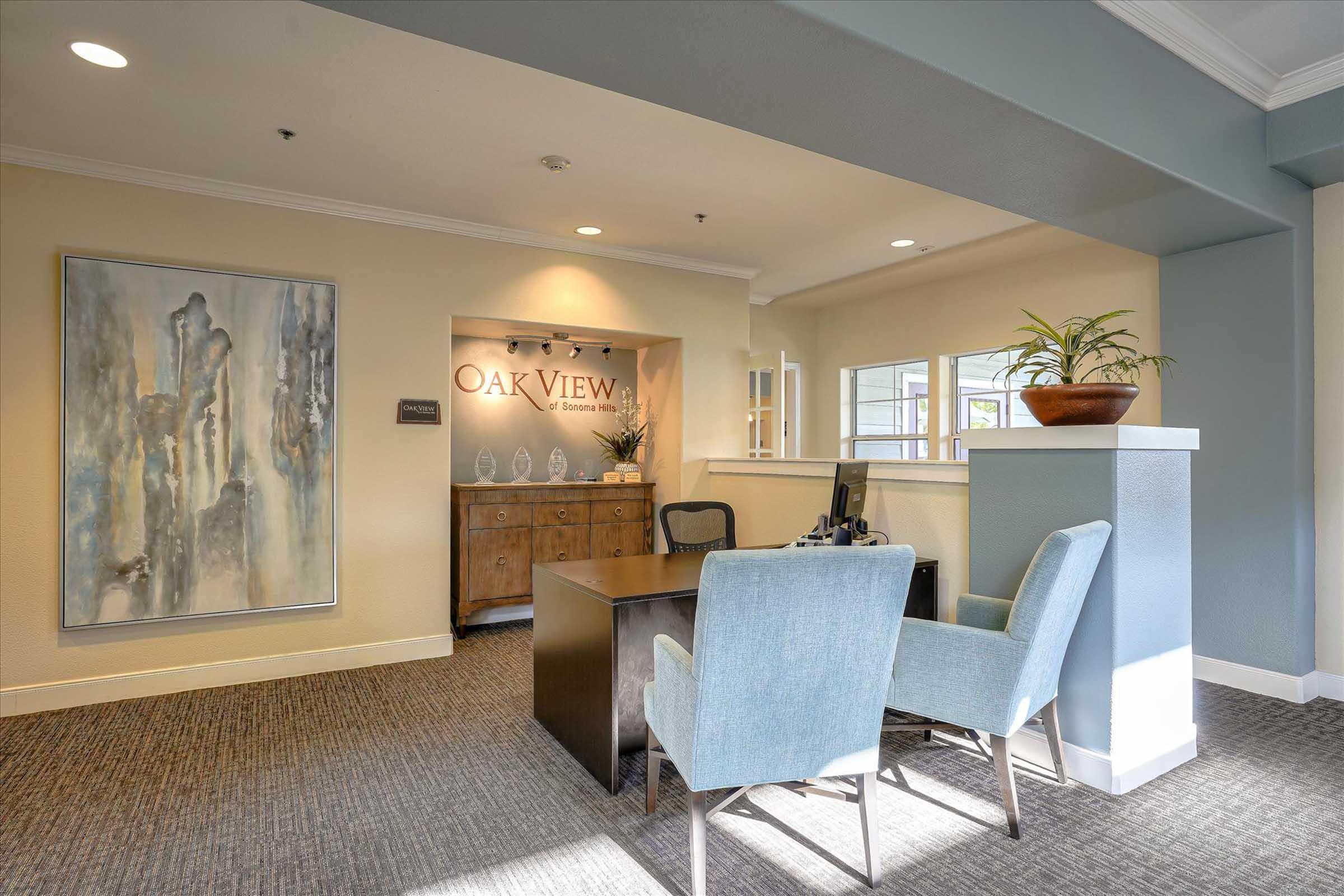 Interior of an office reception area featuring a desk with a chair, a decorative plant, and a large artwork on the wall. The backdrop displays the name "Oak View." The space has a warm, inviting atmosphere with natural light coming from nearby windows.