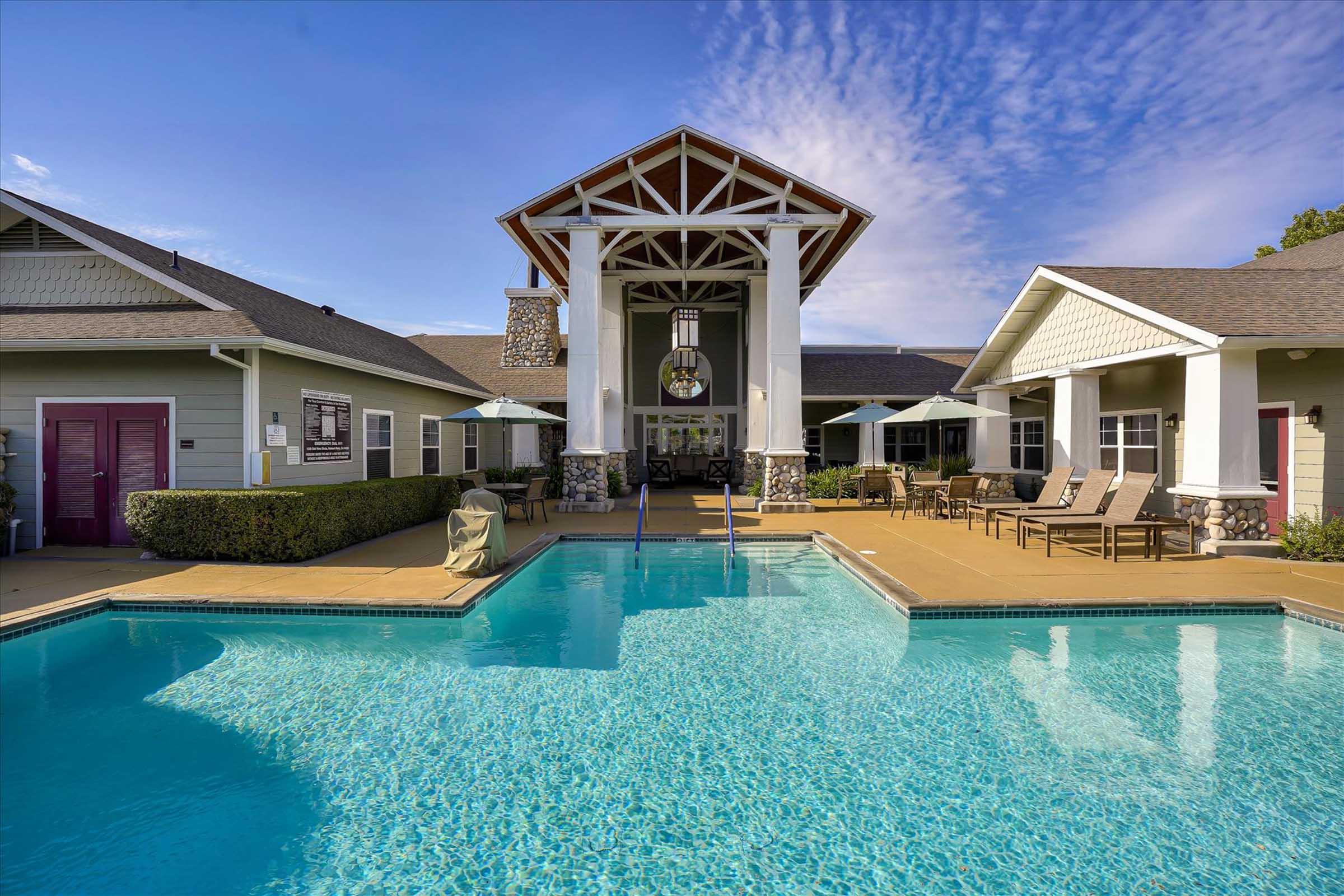 A spacious outdoor pool area with a large swimming pool, lounge chairs, and a nearby clubhouse. The clubhouse features a high ceiling and large windows, surrounded by well-kept landscaping and umbrellas for shade. The sky is clear with a few clouds, creating a serene atmosphere.