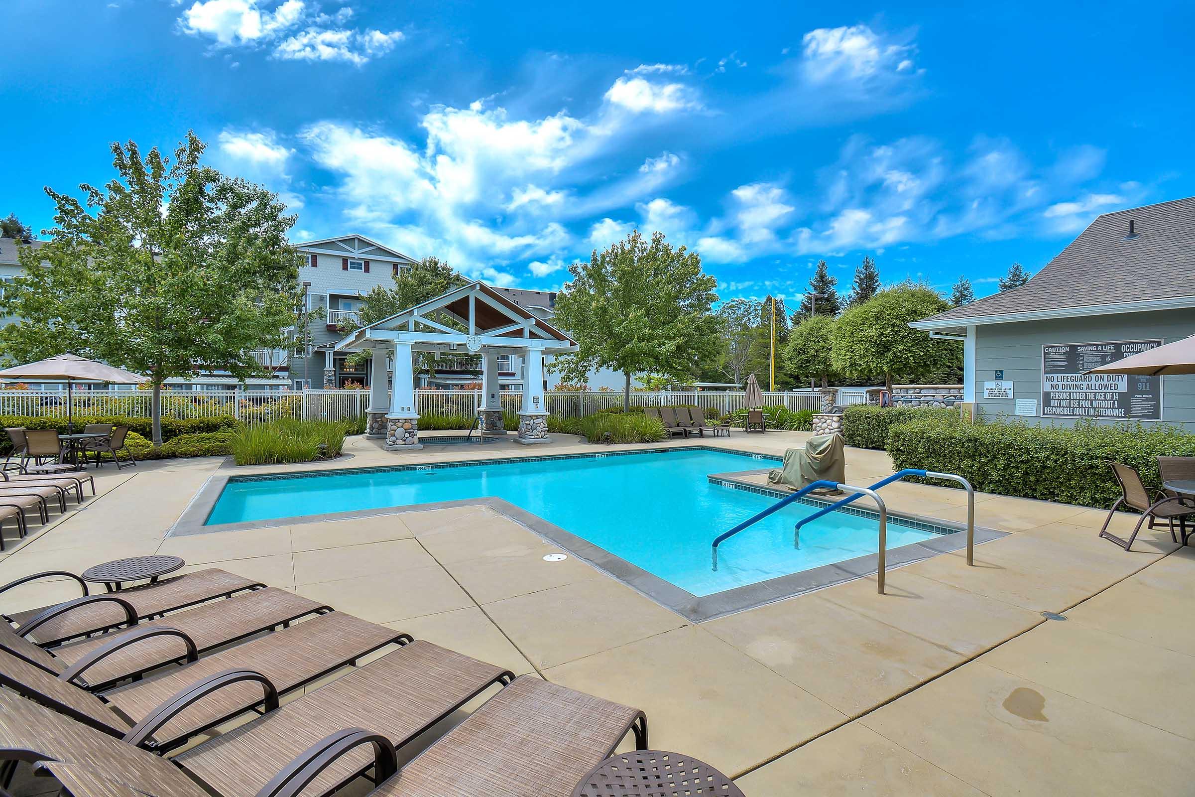 A clear blue swimming pool surrounded by lounge chairs and green landscaping. There is a shaded gazebo in the background, with umbrellas and modern apartment buildings visible. The sky is bright with a few fluffy clouds, creating a serene outdoor atmosphere.
