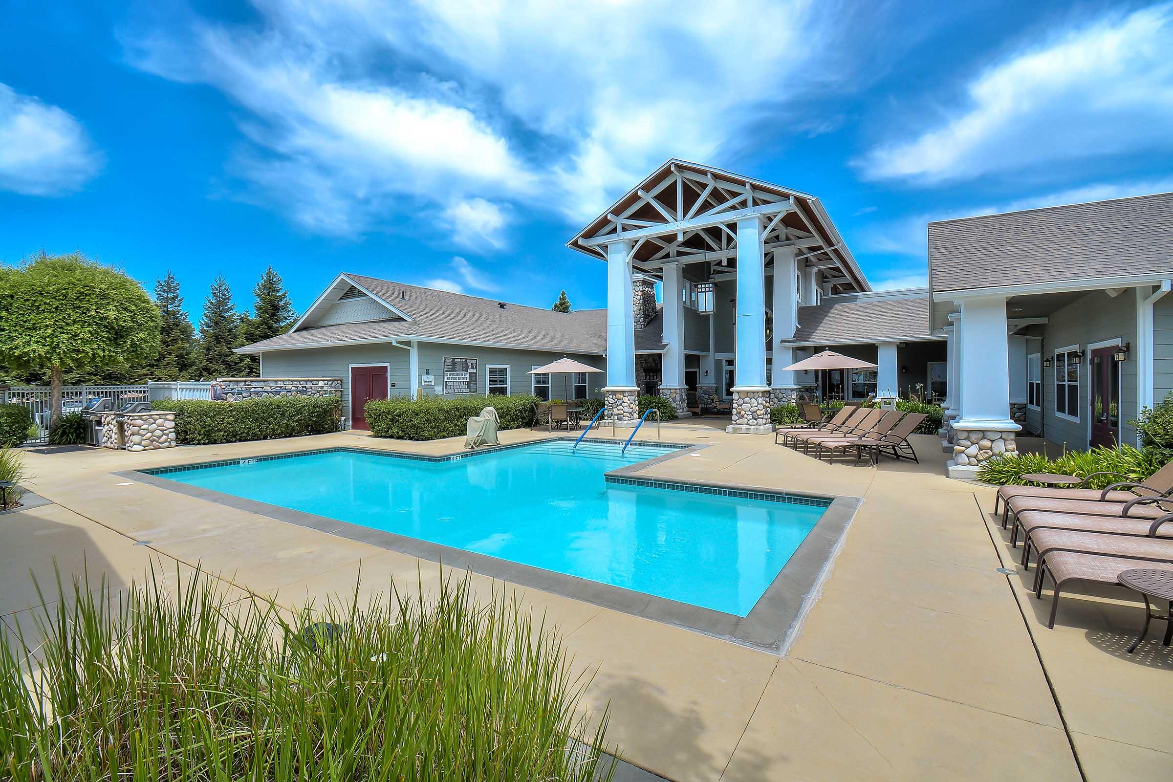 A clear blue swimming pool surrounded by lounge chairs and greenery, located in a well-maintained residential area. The backdrop features a stylish building with a high roof and large windows under a bright blue sky with scattered clouds.