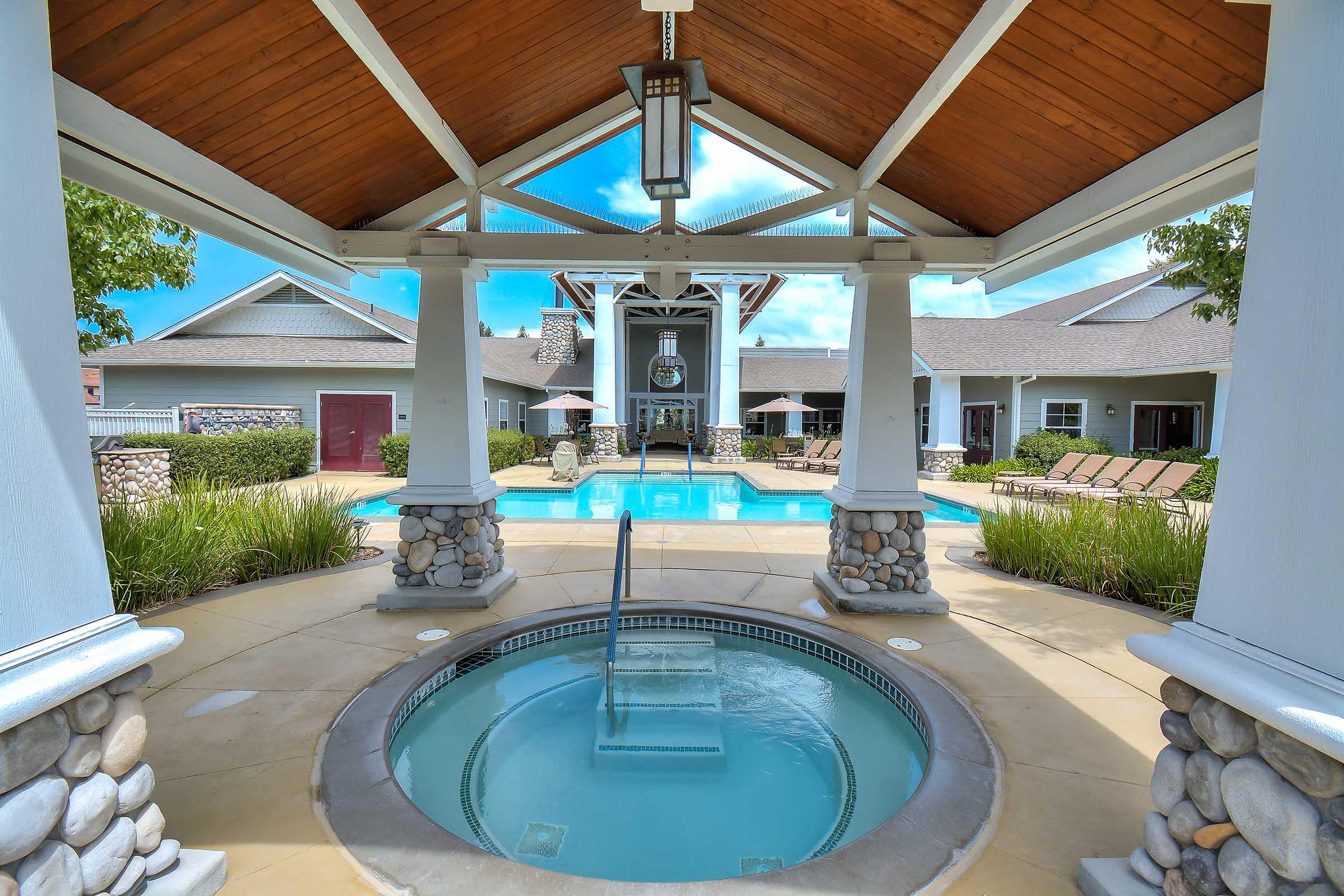 View of a luxurious outdoor pool area featuring a hot tub in the foreground. The pool is surrounded by comfortable lounge chairs, and the backdrop includes a stylish building with a spacious covered area. Lush greenery adds to the relaxing atmosphere under a bright blue sky.