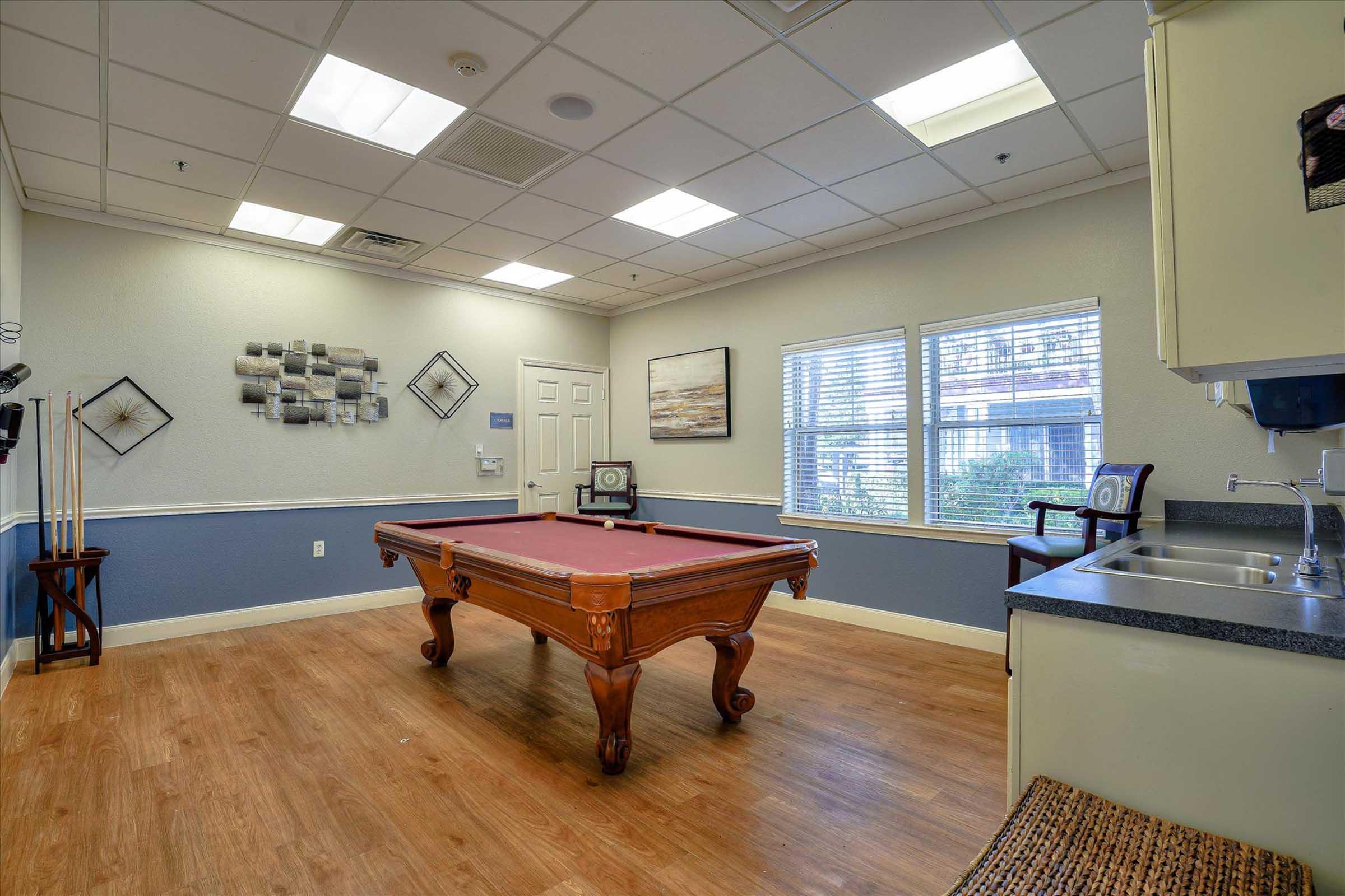A spacious room featuring a billiards table with a rich wooden finish, framed artwork on the walls, a small kitchenette in the corner, and large windows allowing natural light. The flooring is wooden, contributing to a warm and inviting atmosphere.