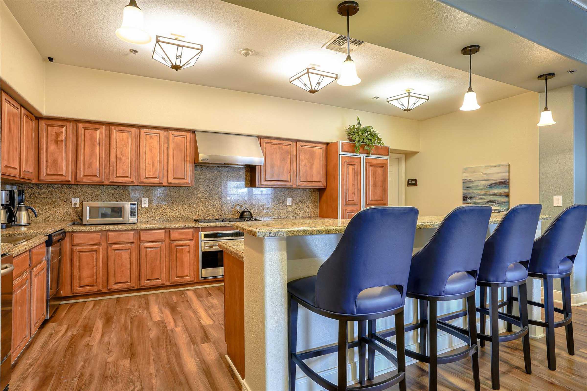 A modern kitchen with wooden cabinets and granite countertops. It features a stainless steel oven and microwave, a sink, and four blue high-backed bar stools at the counter. The space is well-lit with pendant lights and has a decorative painting on the wall.