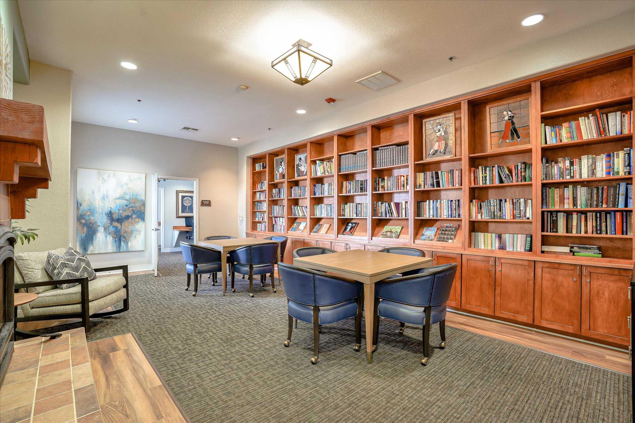 Cozy interior of a library or reading area featuring built-in bookshelves filled with books, two wooden tables surrounded by blue chairs, and a fireplace. Natural light enters the room, creating a welcoming atmosphere for reading and study.