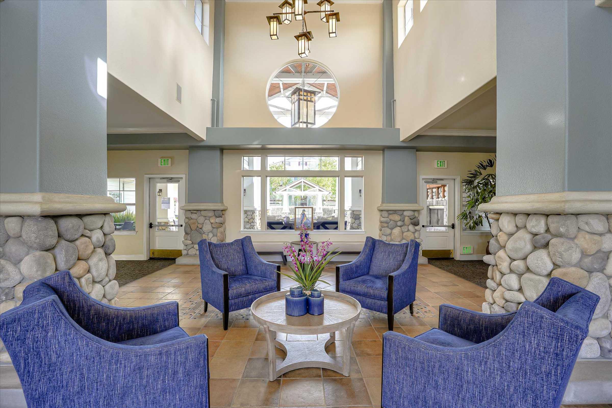 A spacious lobby area featuring four blue upholstered chairs arranged around a round table with a floral centerpiece. Natural light streams in through large windows, highlighting the stone walls and elegant light fixtures. The decor is modern and welcoming, creating a comfortable atmosphere.