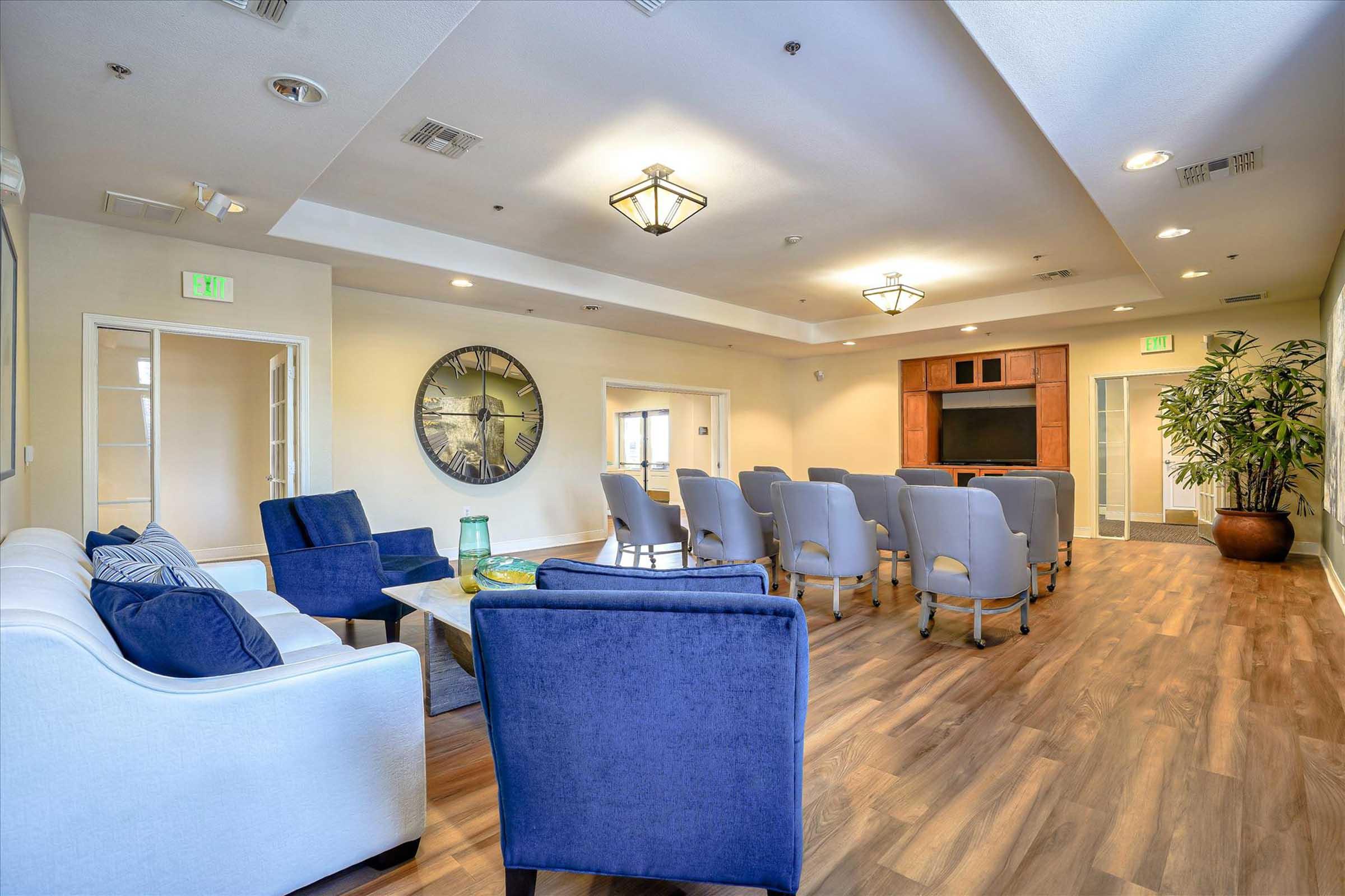 A spacious communal room featuring a comfortable seating arrangement with blue and white chairs, a large wall clock, and a television setup. The room is well-lit with ceiling lights, has wooden flooring, and includes a potted plant for added decor.