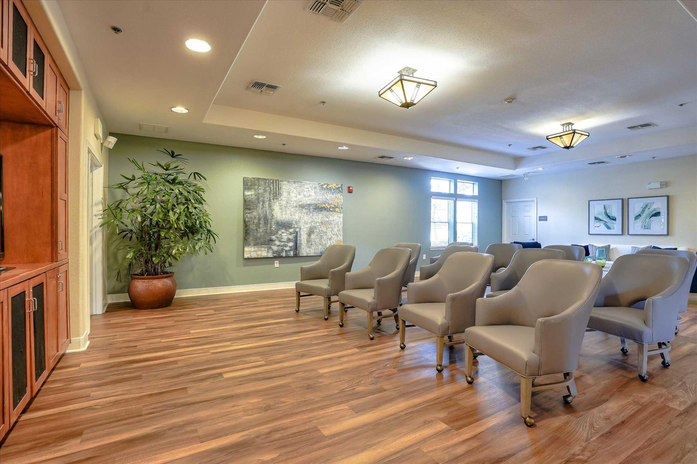 A well-lit waiting area with hardwood floors, featuring several gray armchairs arranged neatly. To one side, there is a large potted plant. A wall holds an abstract painting, and decorative ceiling lights illuminate the space. The overall atmosphere is modern and welcoming.
