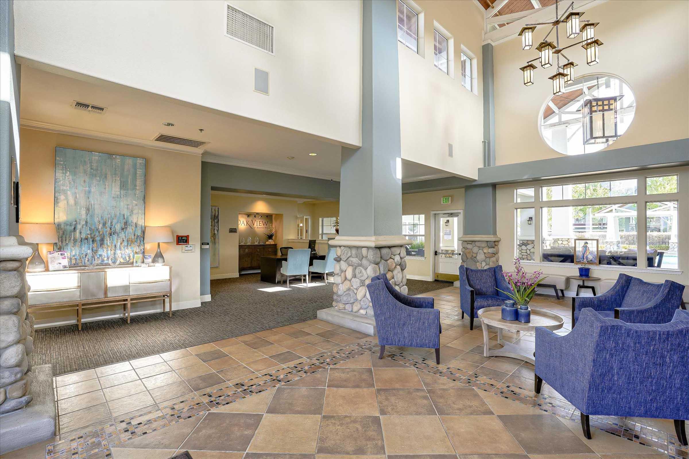 A spacious and modern lobby with high ceilings, featuring a cozy seating area with blue chairs, a decorative stone wall, and a circular window. The floor is tiled with an attractive pattern, and artwork hangs on the wall. Bright lighting and large windows create an inviting atmosphere.