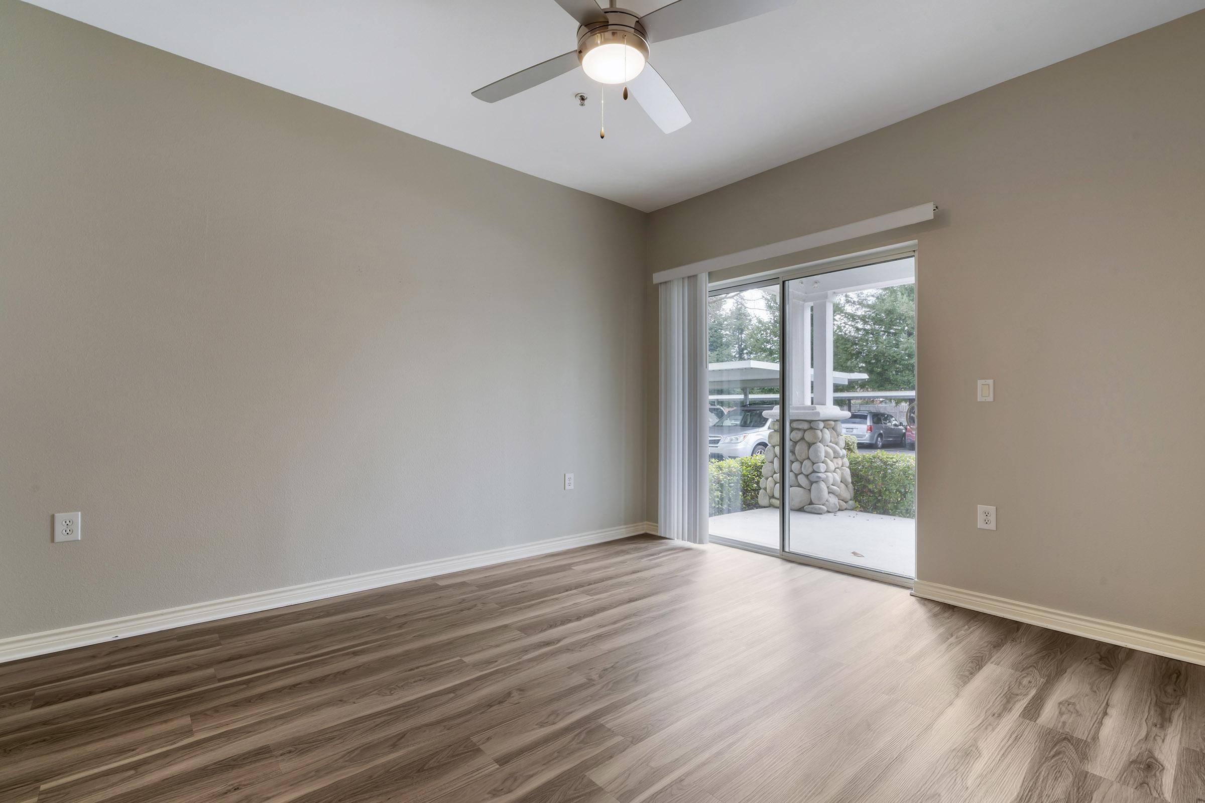Empty room with light beige walls and a ceiling fan. The floor is made of light wood, and there is a large sliding glass door leading to an outdoor view. Natural light is entering through the door, illuminating the space.