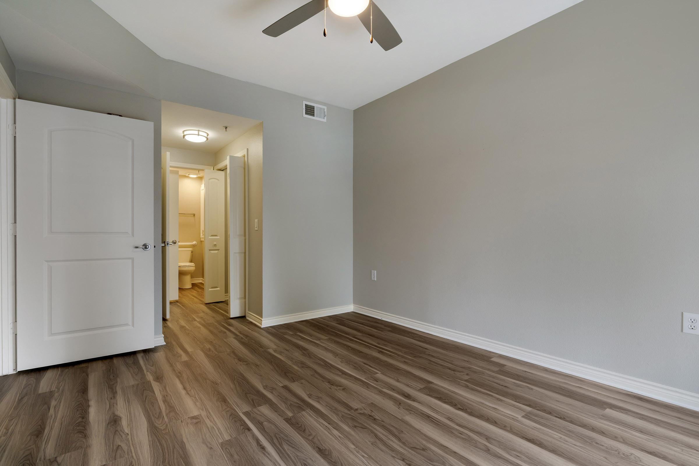 An empty room with a light gray wall and new wood-like flooring. A ceiling fan hangs above, and there are two white doors; one leads to a bathroom visible in the background. The space is bright and uncluttered, showcasing its potential for decoration and furniture placement.