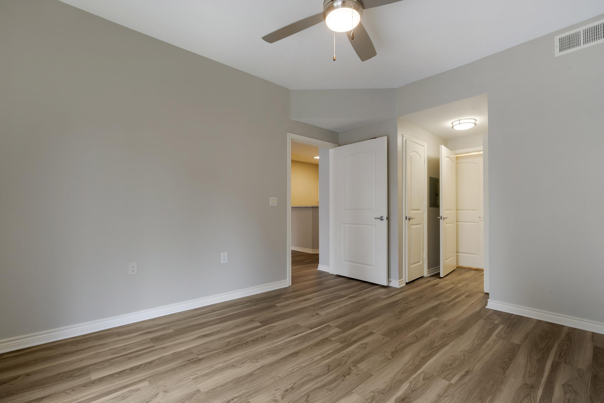 A spacious, empty room with light gray walls and a smooth wooden floor. A ceiling fan is mounted overhead. Two doorways lead to adjacent areas, and soft, natural light enters through the openings, creating a bright and airy atmosphere.