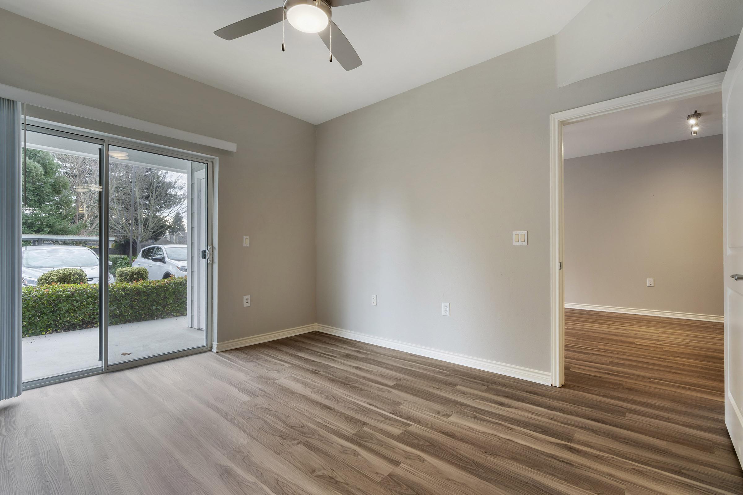 A spacious room with light gray walls and wooden flooring, featuring a ceiling fan. One wall has a sliding glass door leading to an outdoor area, while another wall has an open doorway. The room is well-lit with natural light and has a minimalist design.