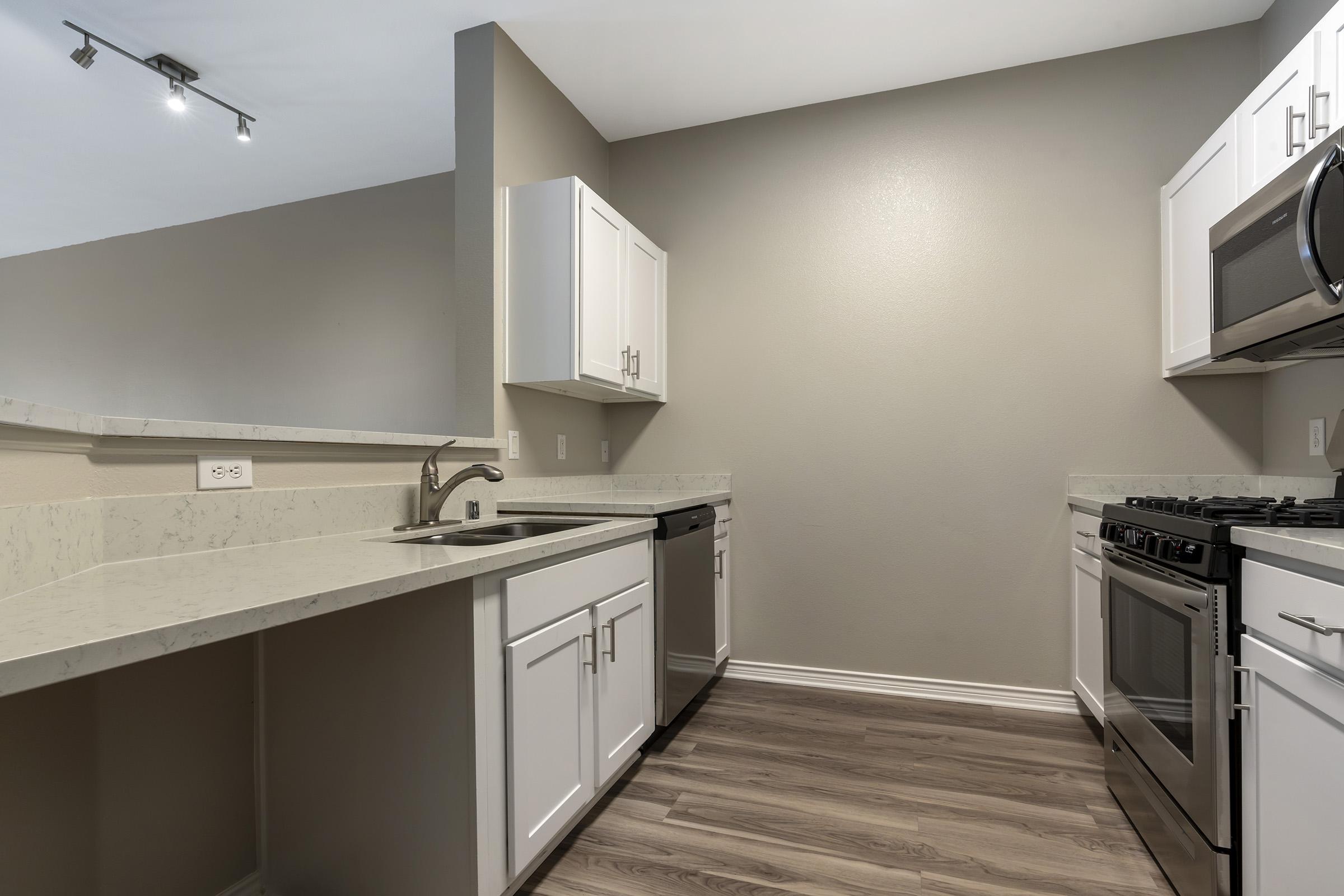 Modern kitchen featuring white cabinets, stainless steel appliances, and a light countertops. The space includes a sink, dishwasher, and an oven, with wood-style flooring and neutral wall colors, creating a clean and inviting atmosphere. Ceiling lights provide additional illumination.