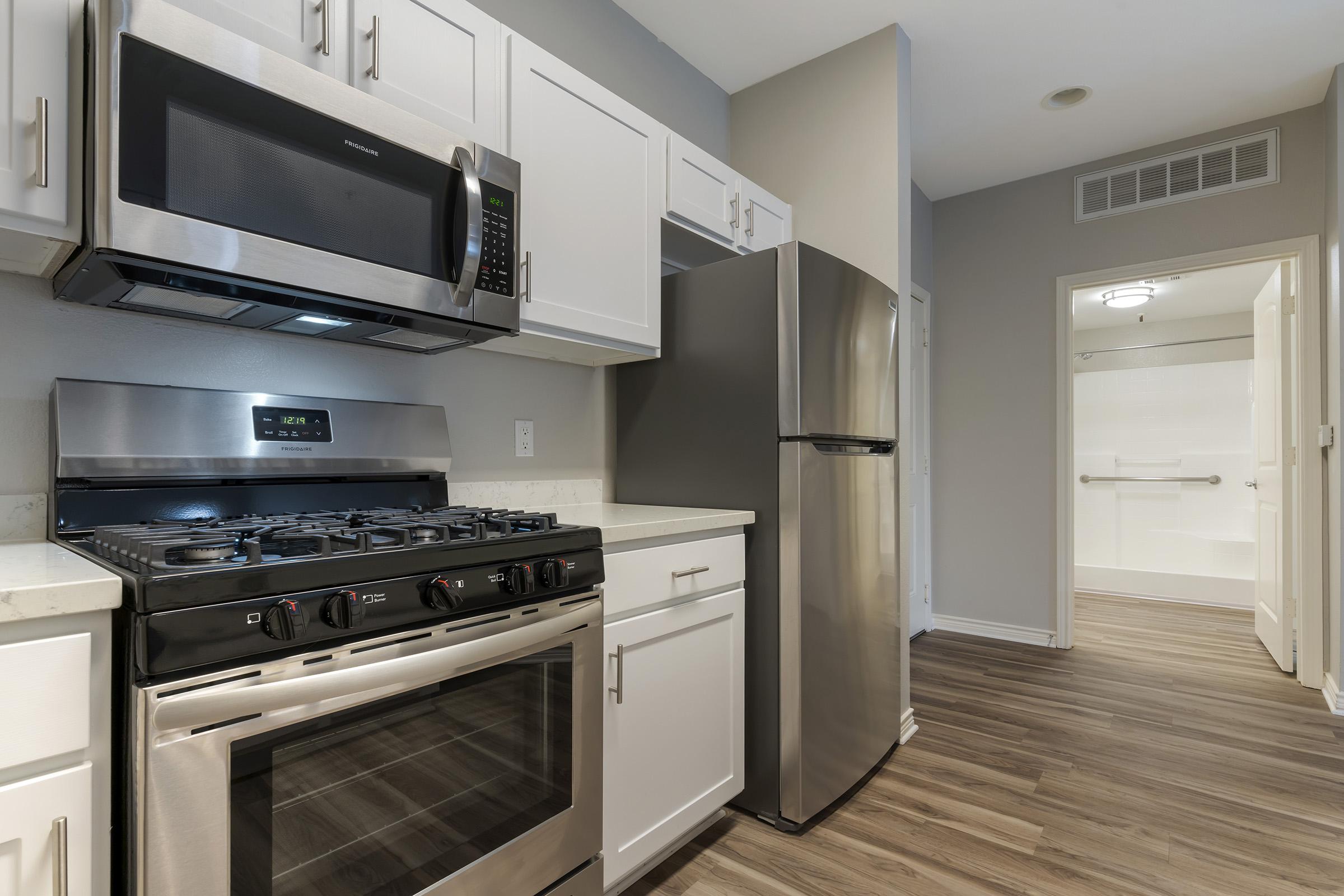 Modern kitchen featuring stainless steel appliances, including a microwave, gas stove, and refrigerator. White cabinetry provides a clean, contemporary look. The floor is covered with wood-like flooring, and there's a doorway leading to another room with minimal natural light.