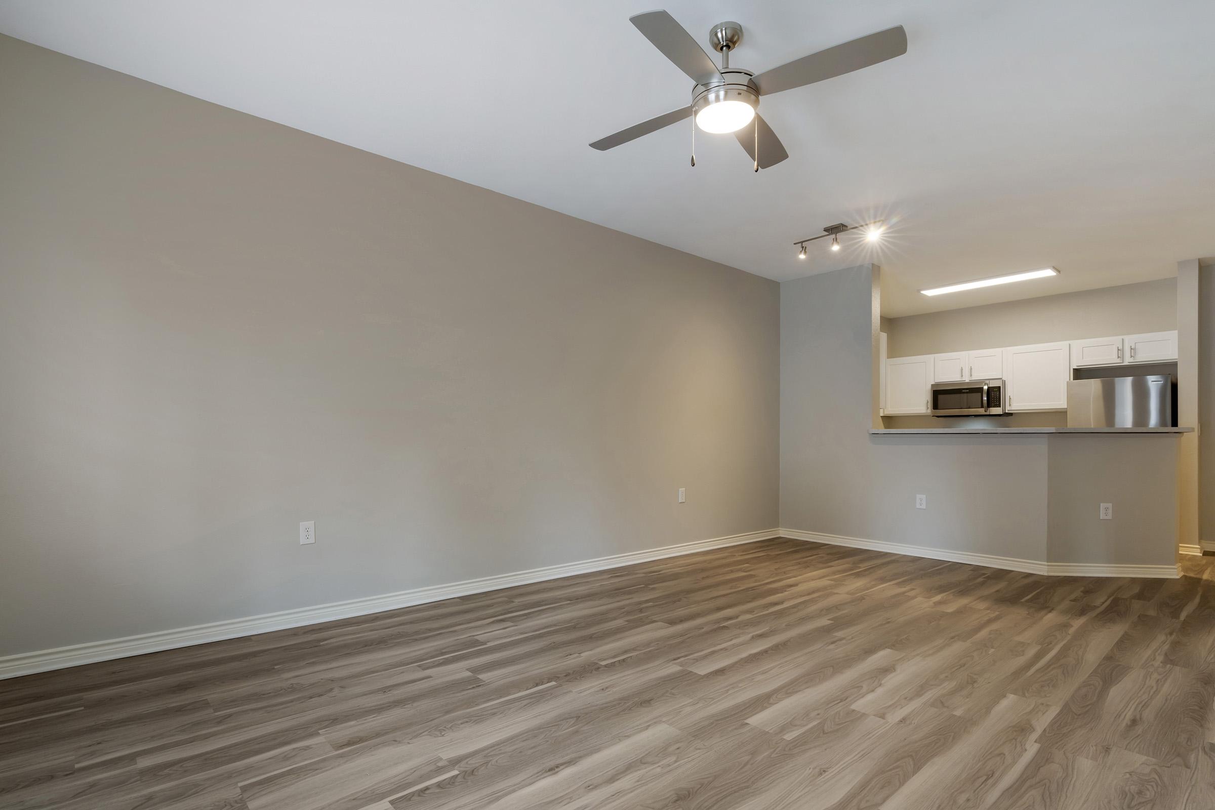 A spacious, modern living area with light-colored walls and wooden flooring. A ceiling fan is installed, and there's a kitchen bar viewable in the background with white cabinetry, a microwave, and a stainless steel refrigerator. The space is well-lit and has a minimalistic design.