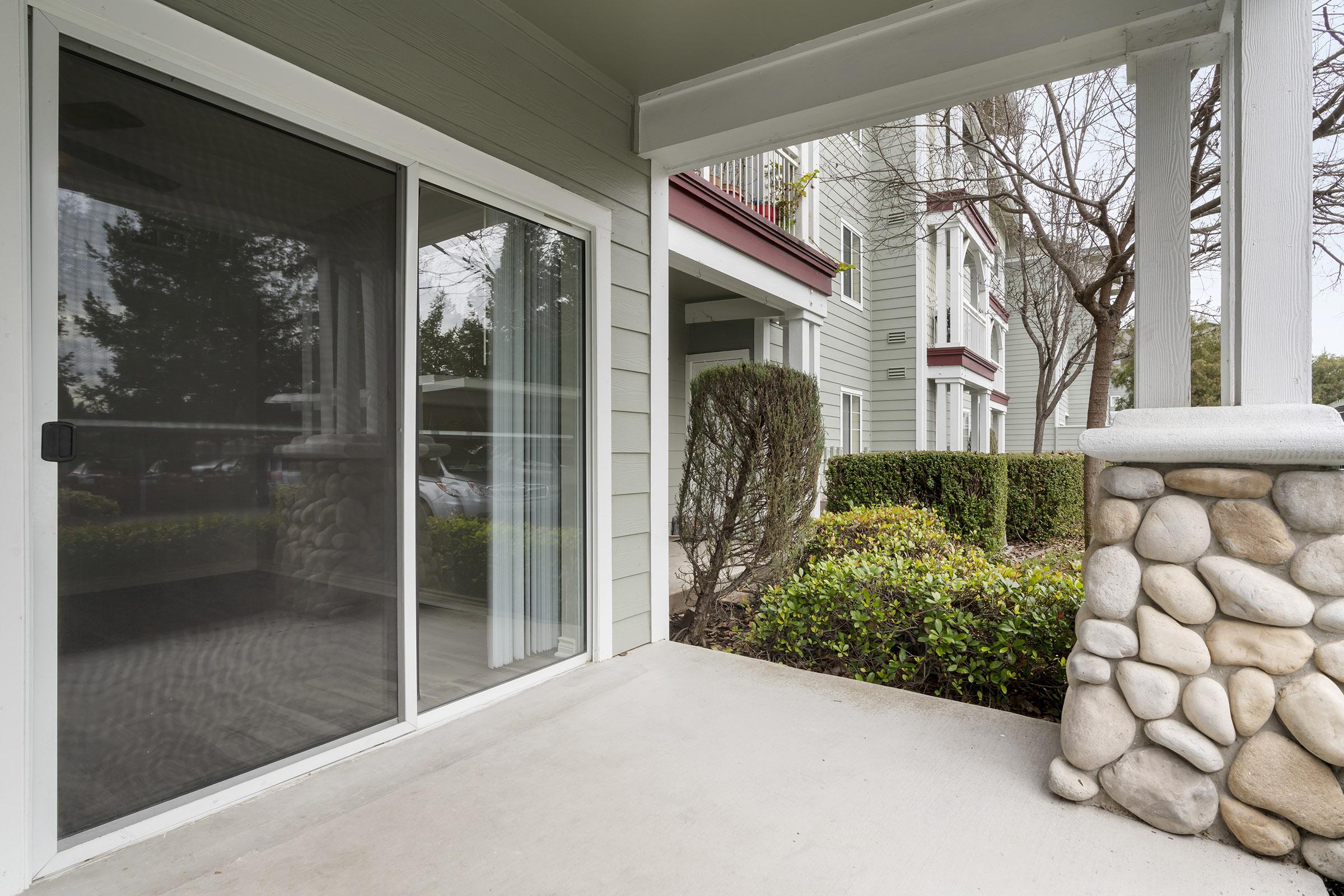 A patio with a sliding glass door, featuring a concrete floor and a stone pillar. In the background, there are shrubs and a multi-story residential building, partially obscured by trees.