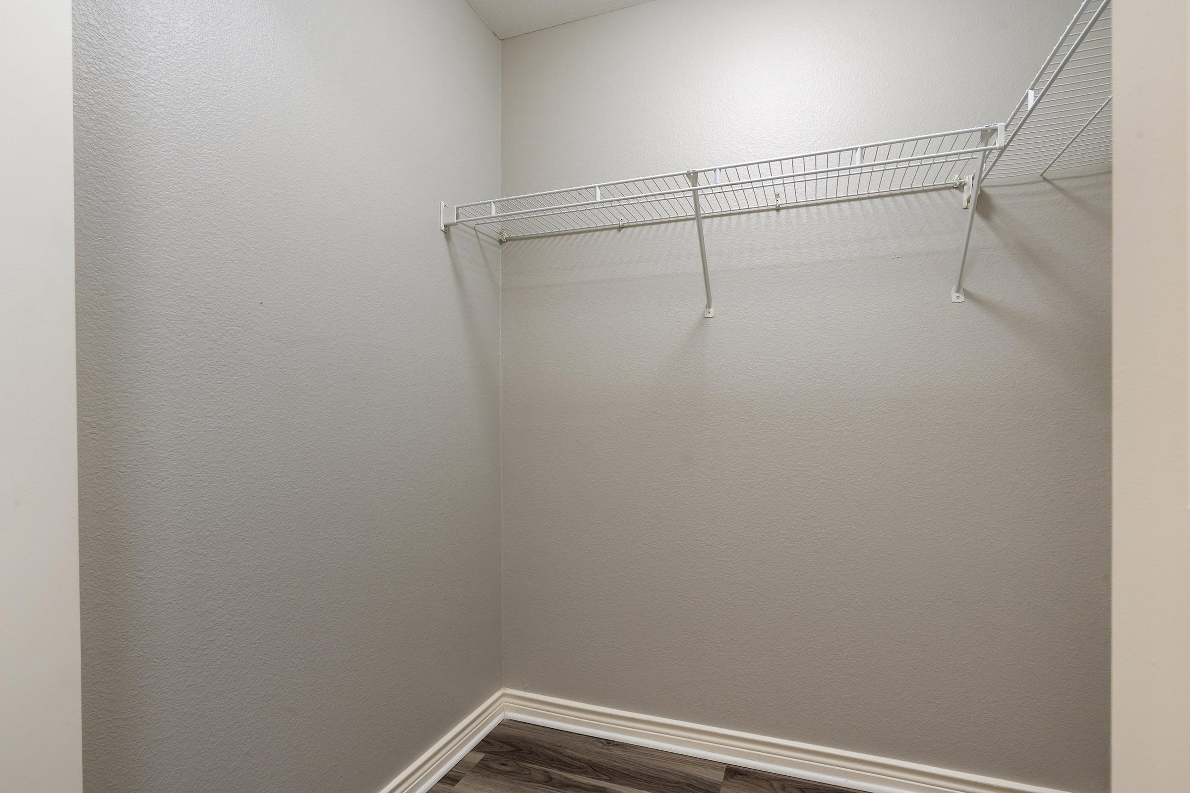Empty closet with plain gray walls and a wire shelf mounted on one side. The floor features light-colored wood-like laminate, creating a clean and minimalistic storage space.