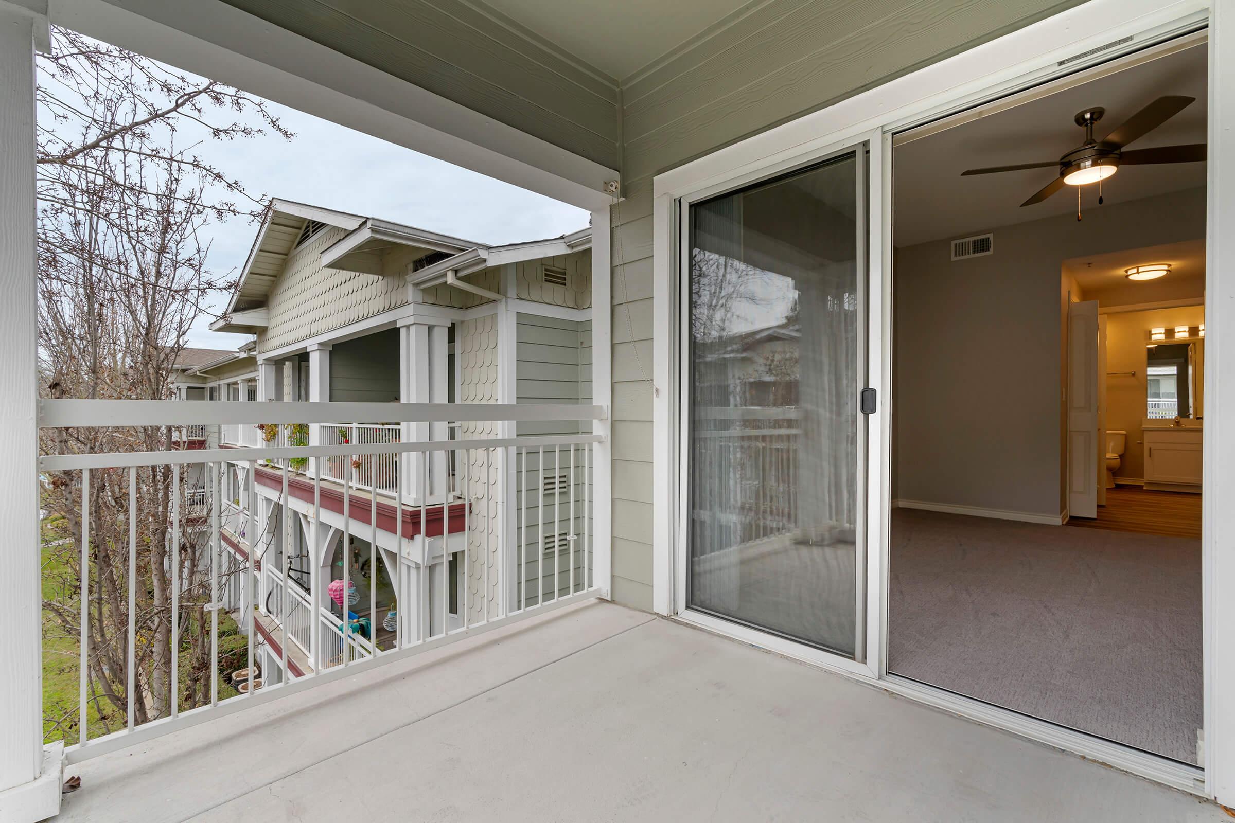 A view from a balcony showing a spacious outdoor area with a sliding glass door leading into an interior room. In the background, neighboring apartment buildings are visible. The scene features a cloudy sky and greenery, creating a tranquil residential environment.