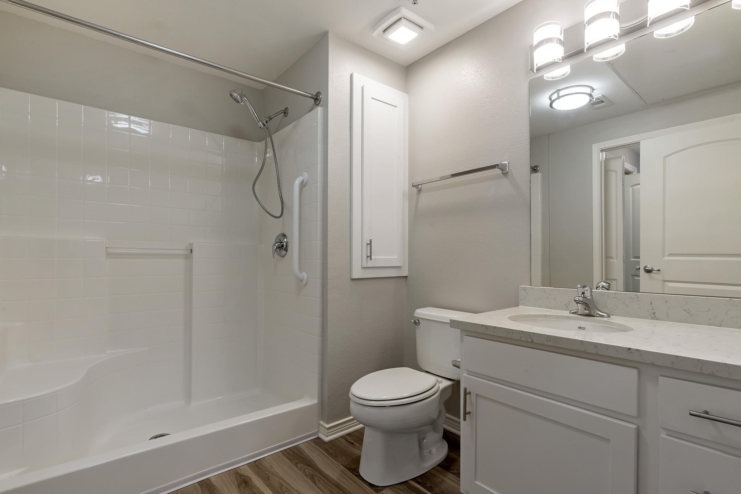 A clean, modern bathroom featuring a white bathtub with a shower, a toilet, and a sink with a countertop. The walls are painted light gray, and there is a mirror above the sink. Bright lighting fixtures are installed on the ceiling, and the floor has a wood-like appearance.