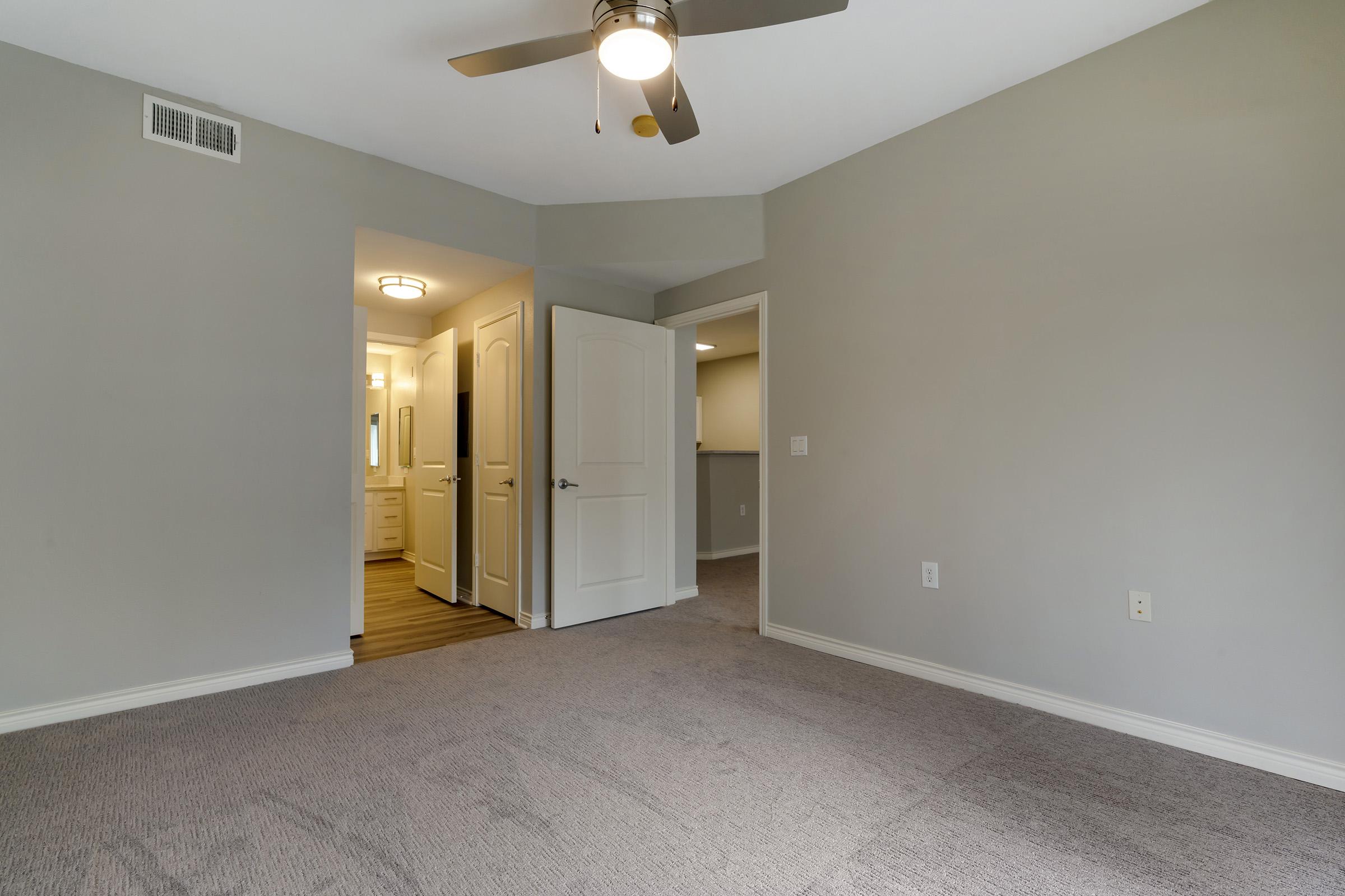 A spacious, carpeted room with light gray walls and a ceiling fan. A doorway leads to a bathroom with modern fixtures, visible in the background. The room features a natural light source and neutral colors, creating a calm and airy atmosphere. The flooring appears modern and inviting.