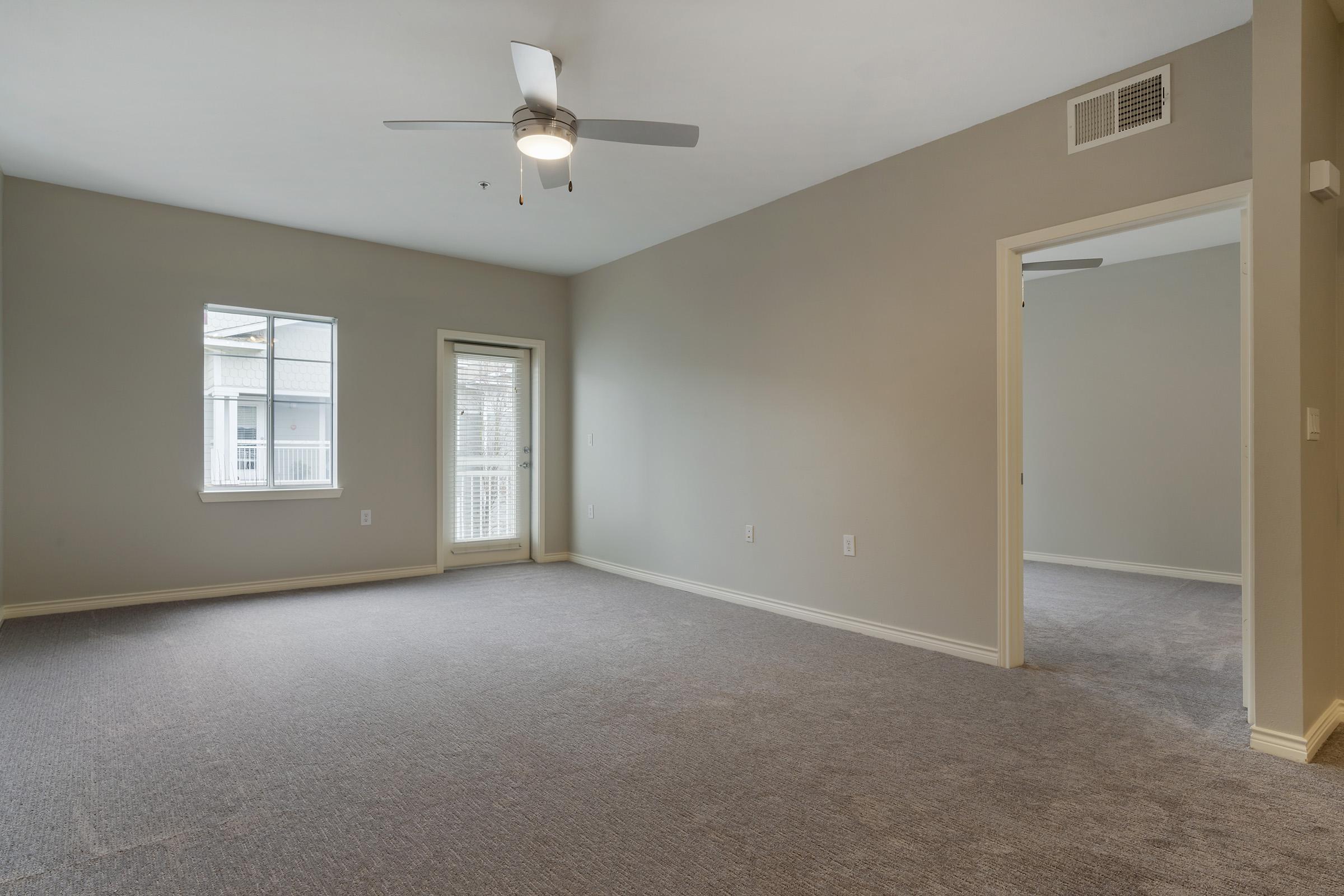 Empty room with gray walls and carpet, featuring a ceiling fan. A window on the left side allows natural light, while a door leads to a balcony. Another doorway on the right connects to an adjacent room.