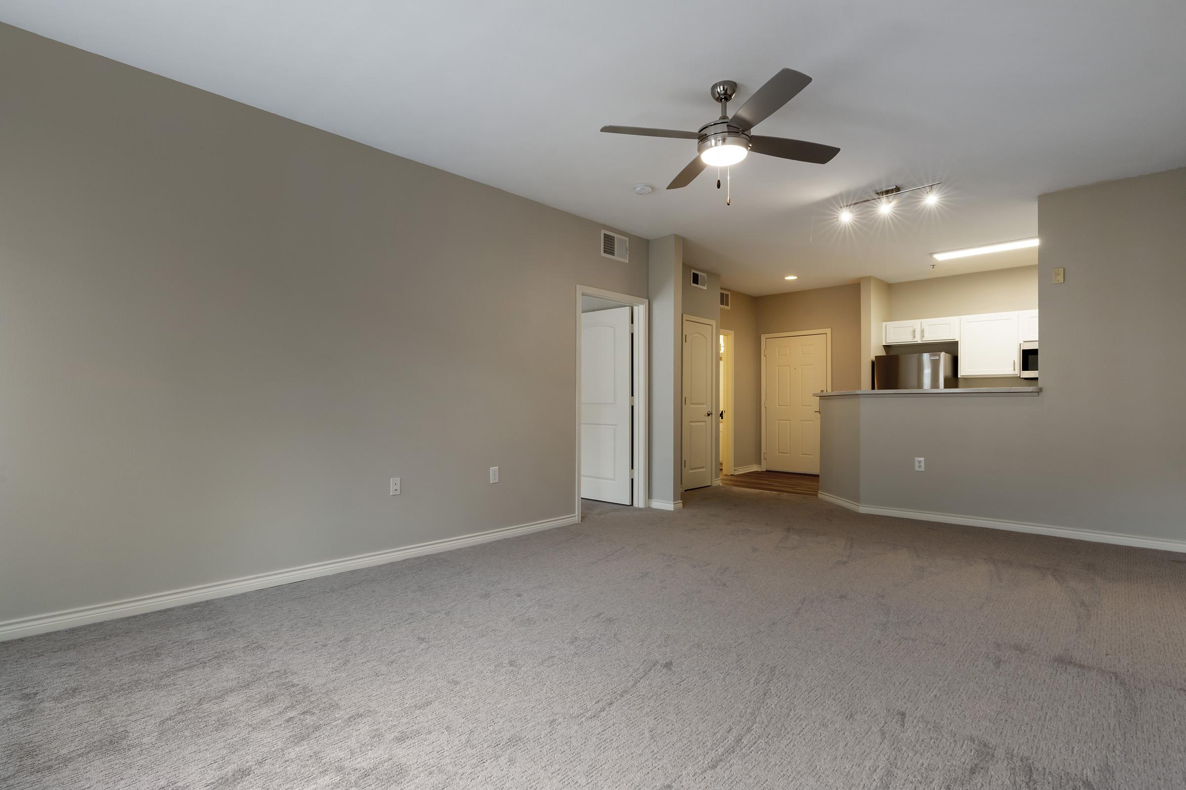 A spacious living area featuring light gray walls and carpet. A ceiling fan is installed, and there's a doorway leading to a kitchen with white cabinets, a refrigerator, and a counter. Soft overhead lighting enhances the open layout, creating a bright and inviting atmosphere.