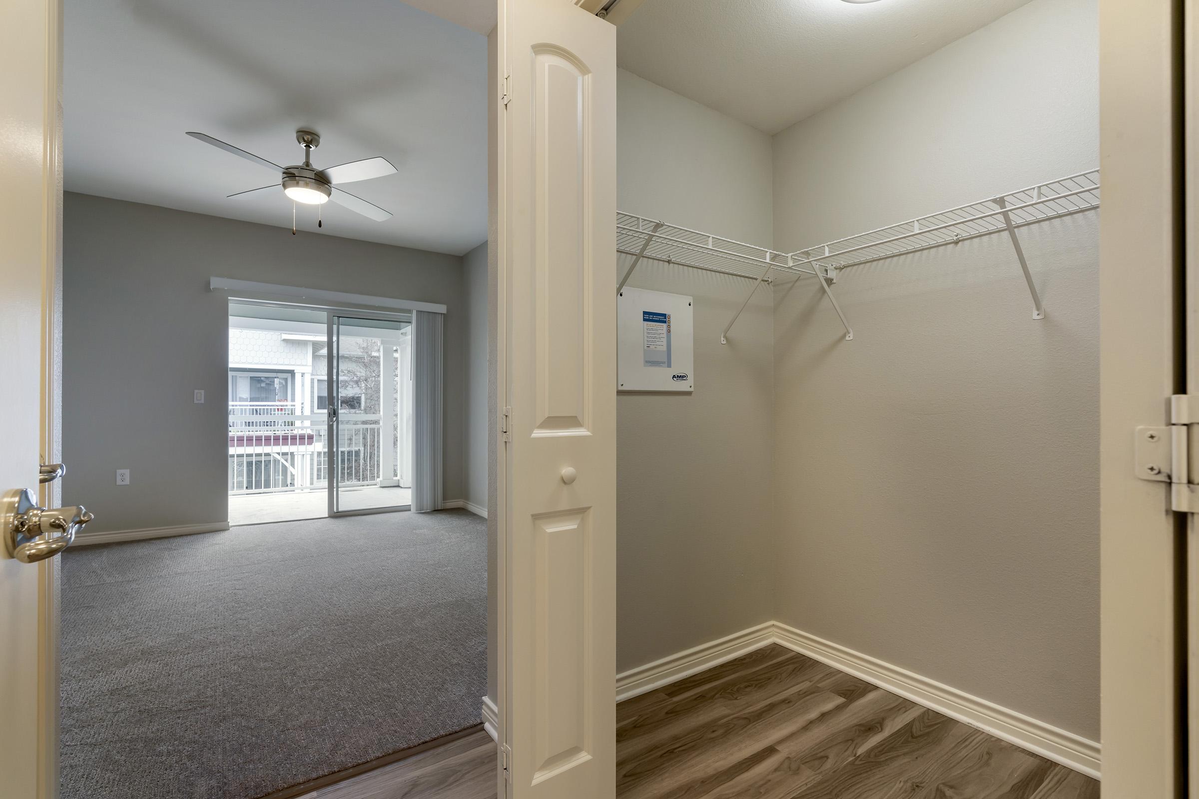 View from an open door into a spacious closet with a hanging rod and a wall hook. Beyond the closet, there's an interior view of a living area featuring a ceiling fan, carpeted flooring, and a sliding glass door leading to an outdoor balcony or patio. The walls are painted in a light color.