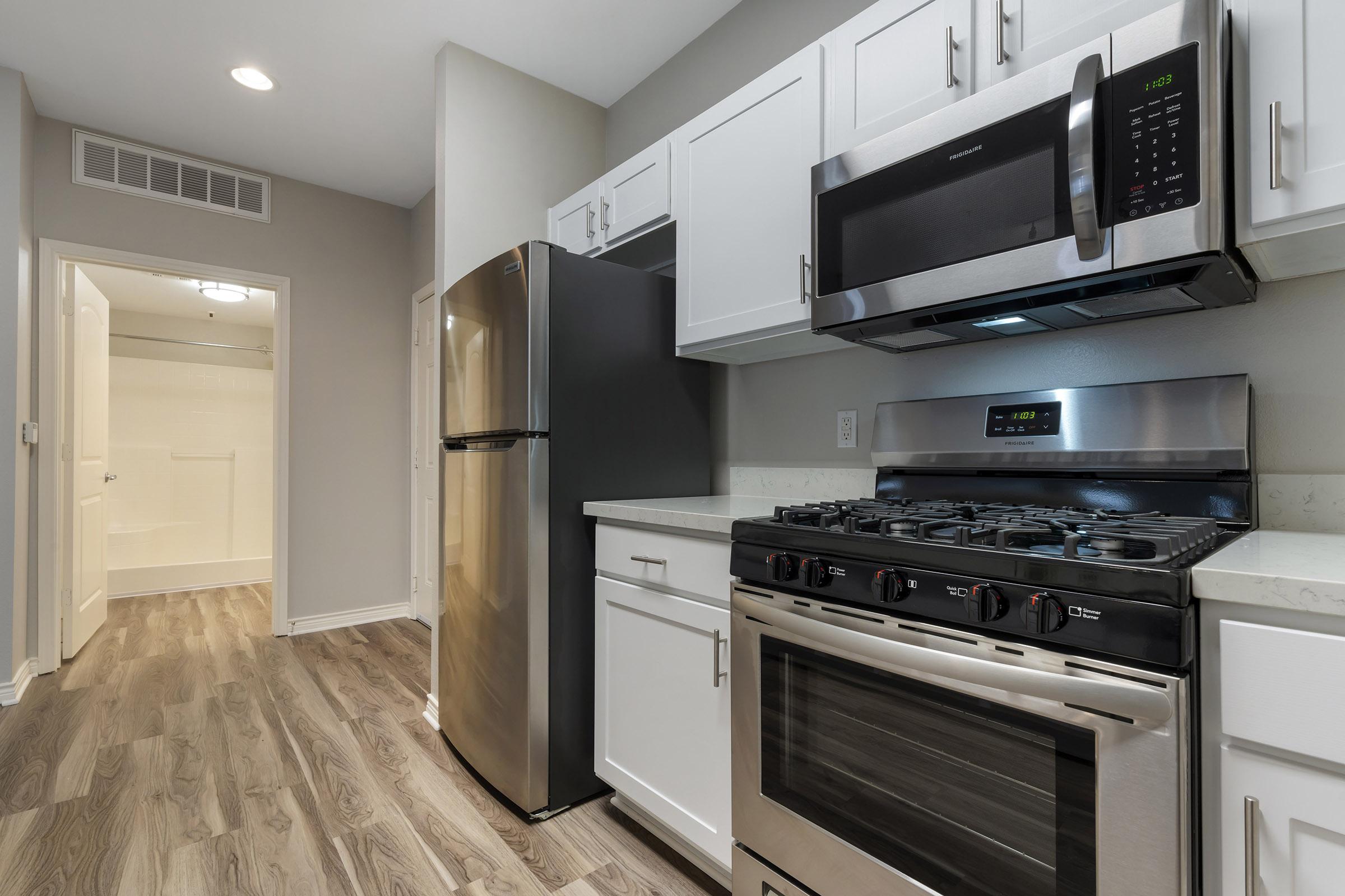 Modern kitchen featuring stainless steel appliances including a gas stove, microwave, and refrigerator. The cabinetry is white with a glossy finish, complemented by a light-colored countertop. The flooring is wood-like laminate. A doorway leads to a bathroom or utility area in the background.