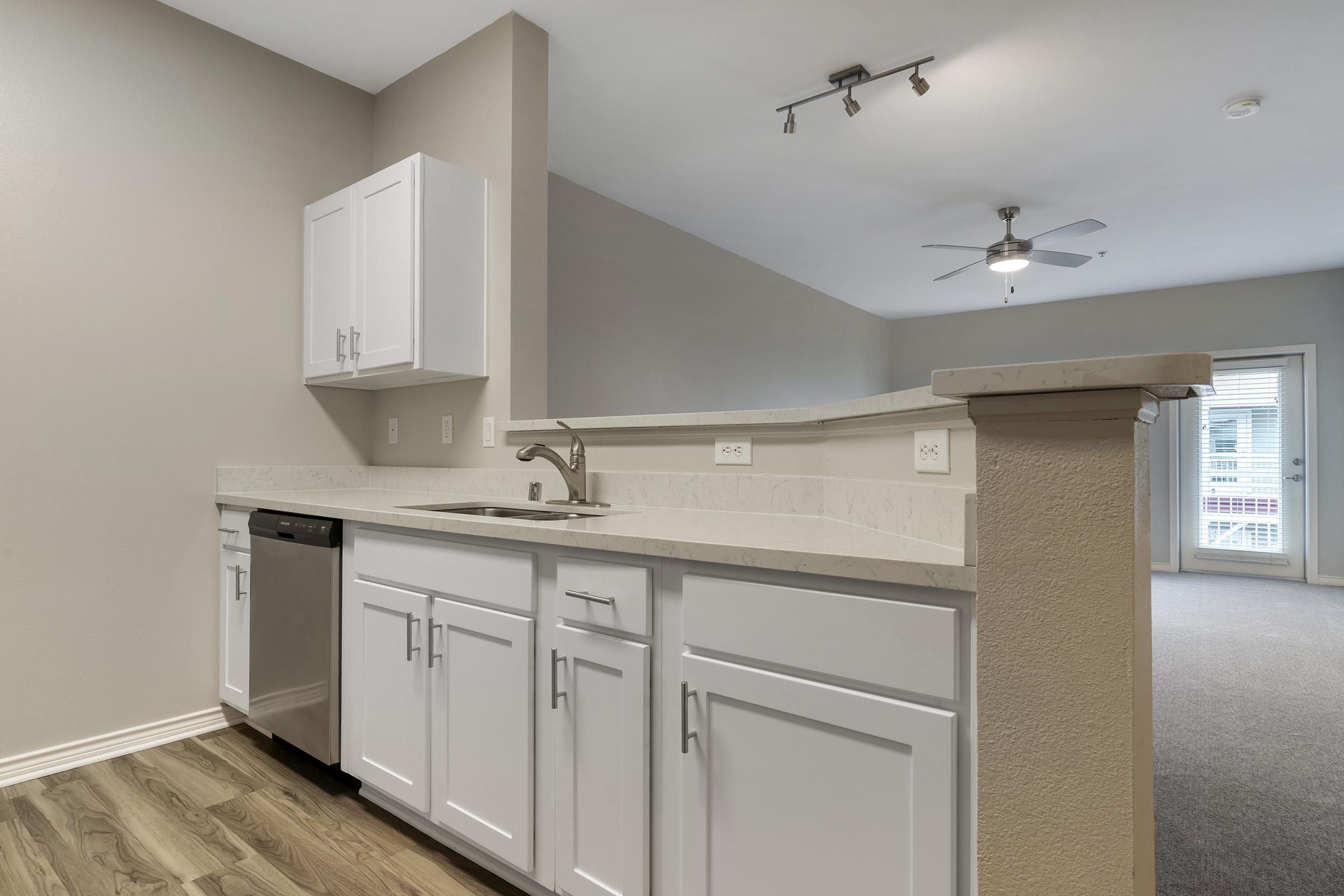 Modern kitchen with white cabinetry, stainless steel dishwasher, and sink, featuring a light-colored countertop. The room has a ceiling fan and is open to a living area with carpet. Neutral walls and ample natural light create a spacious atmosphere.