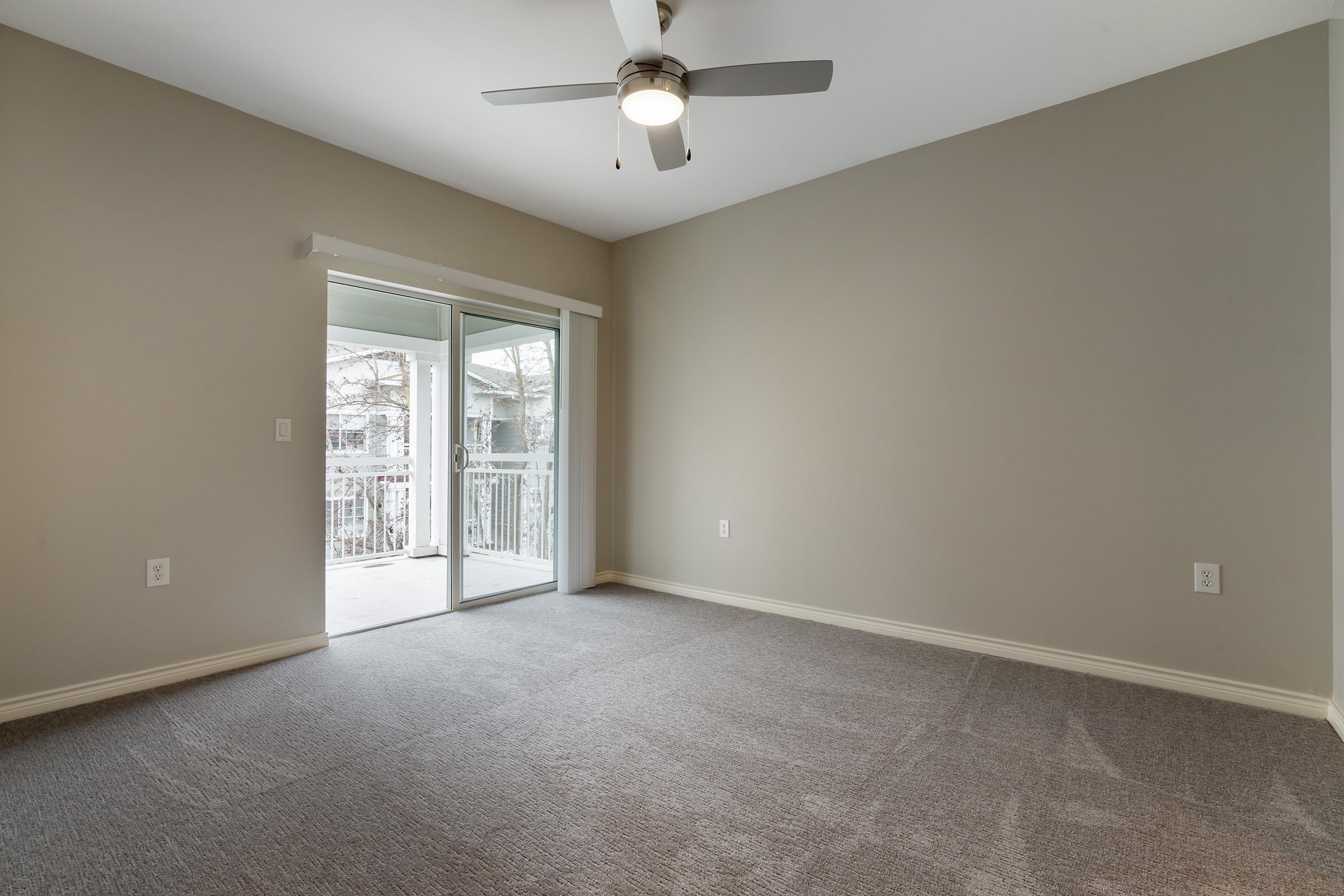 Empty room with light gray walls and carpet. A ceiling fan is mounted in the center, and a sliding glass door leads to a balcony. Natural light fills the space, creating a bright and airy atmosphere. No furnishings are present, emphasizing the room's spaciousness.